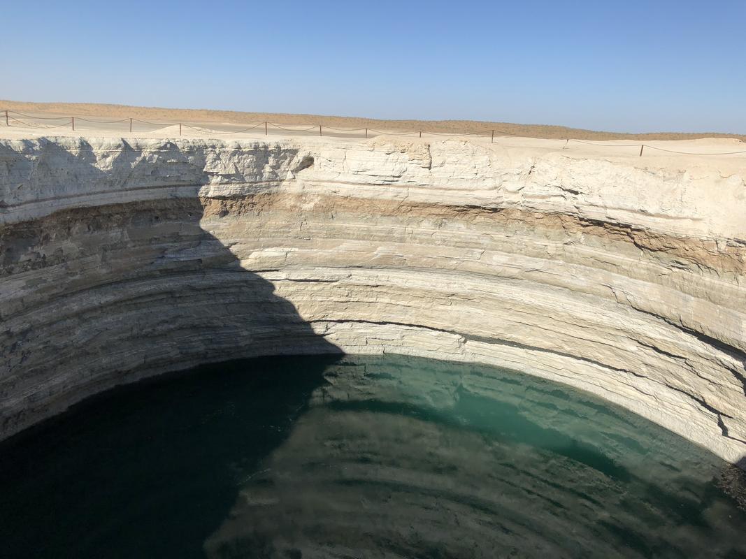 A deep, circular crater with layered rock walls and a pool of blue-green water at its base.