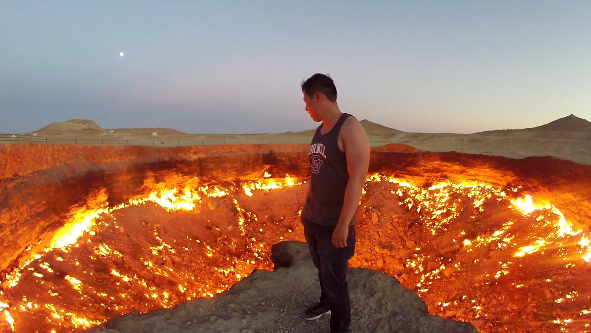 A man stands at the edge of the fiery Darvaza Gas Crater, also known as the Door to Hell, under a twilight sky with the moon visible.