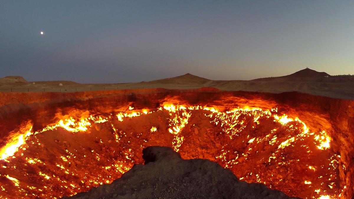 The fiery Darvaza Gas Crater glows under a twilight sky with a visible moon.