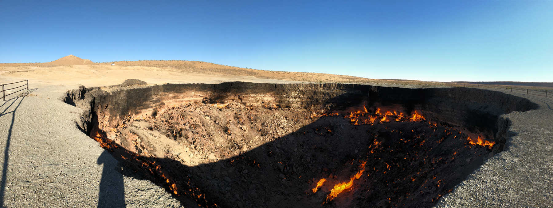 A vast, fiery gas crater burns in the middle of a desert landscape under a clear blue sky.