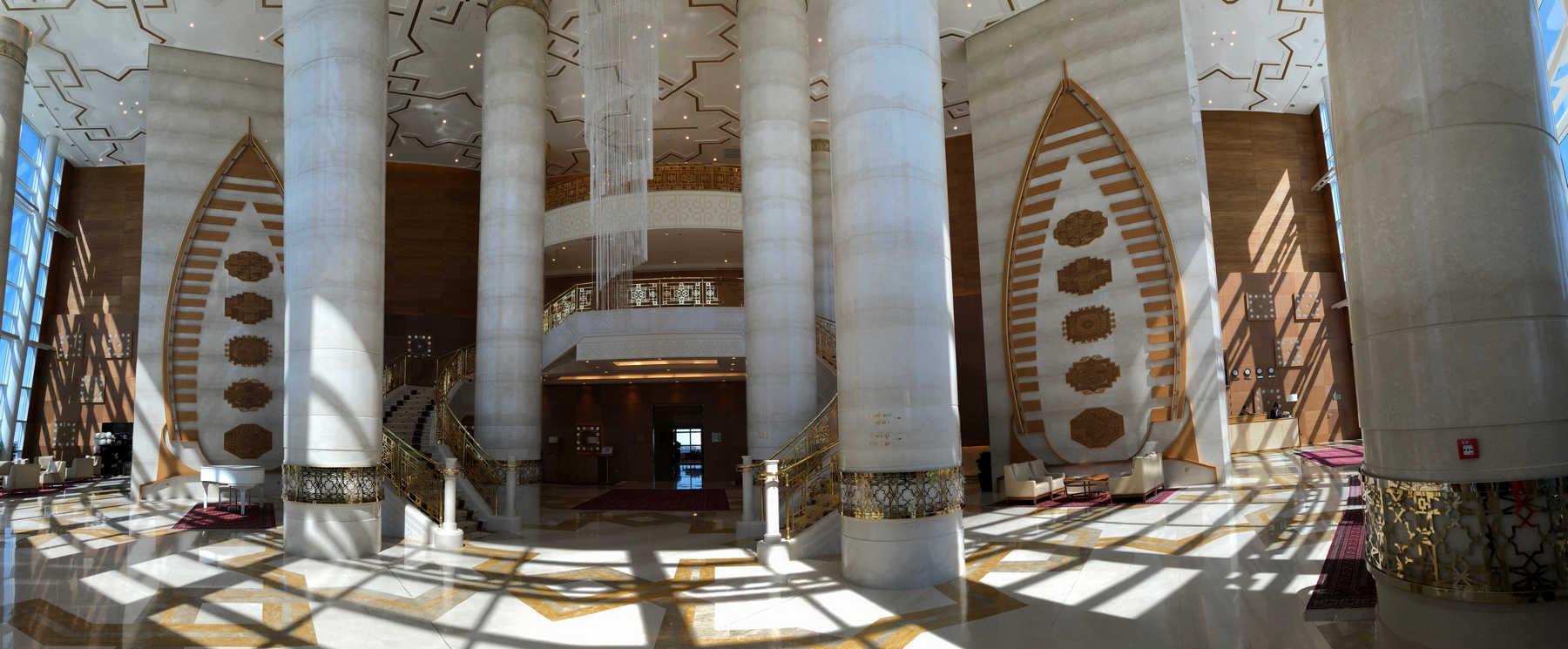 A panoramic view of the opulent Yyldyz Hotel lobby, showcasing towering marble columns, grand staircases, elaborate gold details, and a sparkling chandelier.