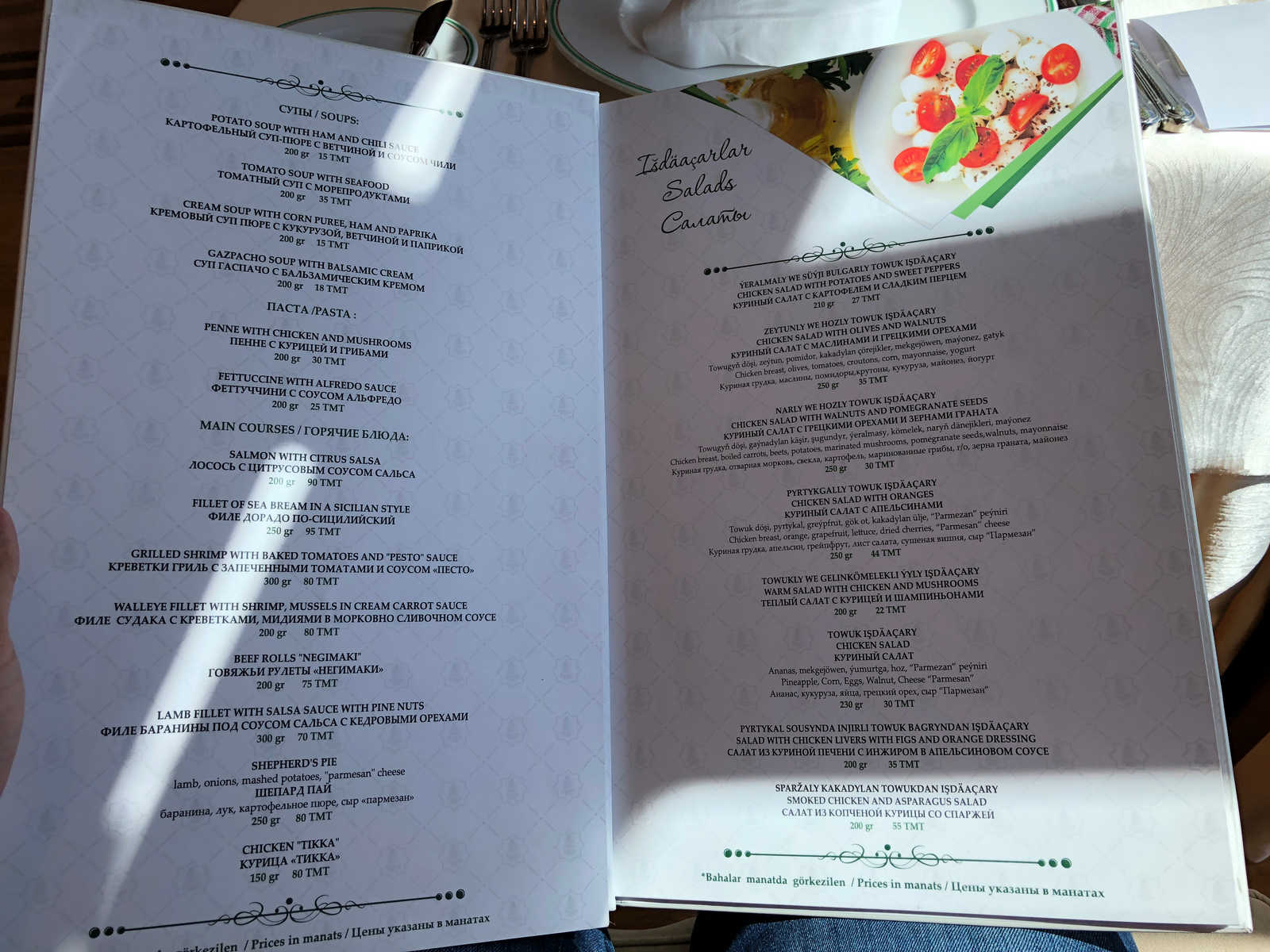 An open restaurant menu displays soups, main courses, and salads, with a picture of a Caprese salad on one page.