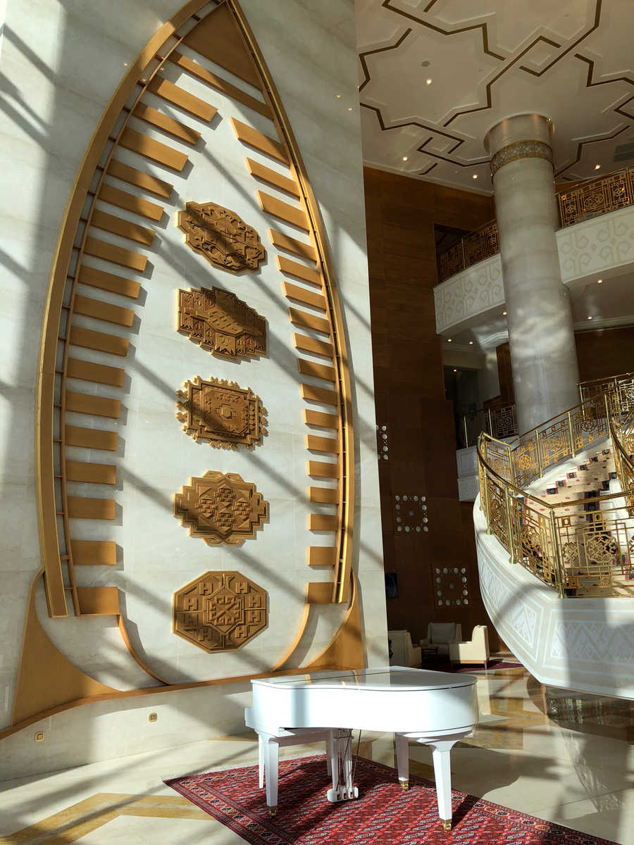The luxurious Yyldyz Hotel lobby features a large boat-shaped wall decoration, a white grand piano, and an ornate curved staircase with gold railings.