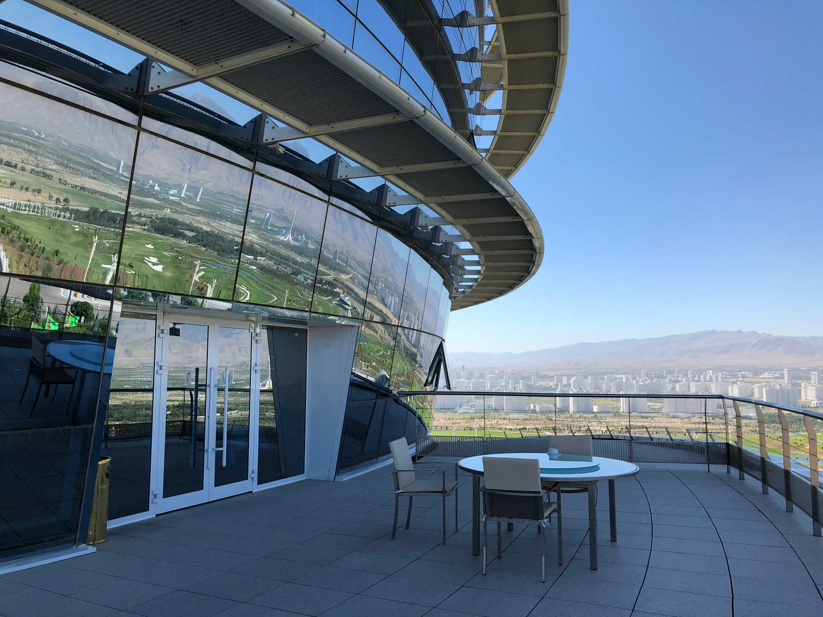 Outdoor terrace of the Yyldyz Hotel with a curved glass facade reflecting a golf course, overlooking a city and mountains.