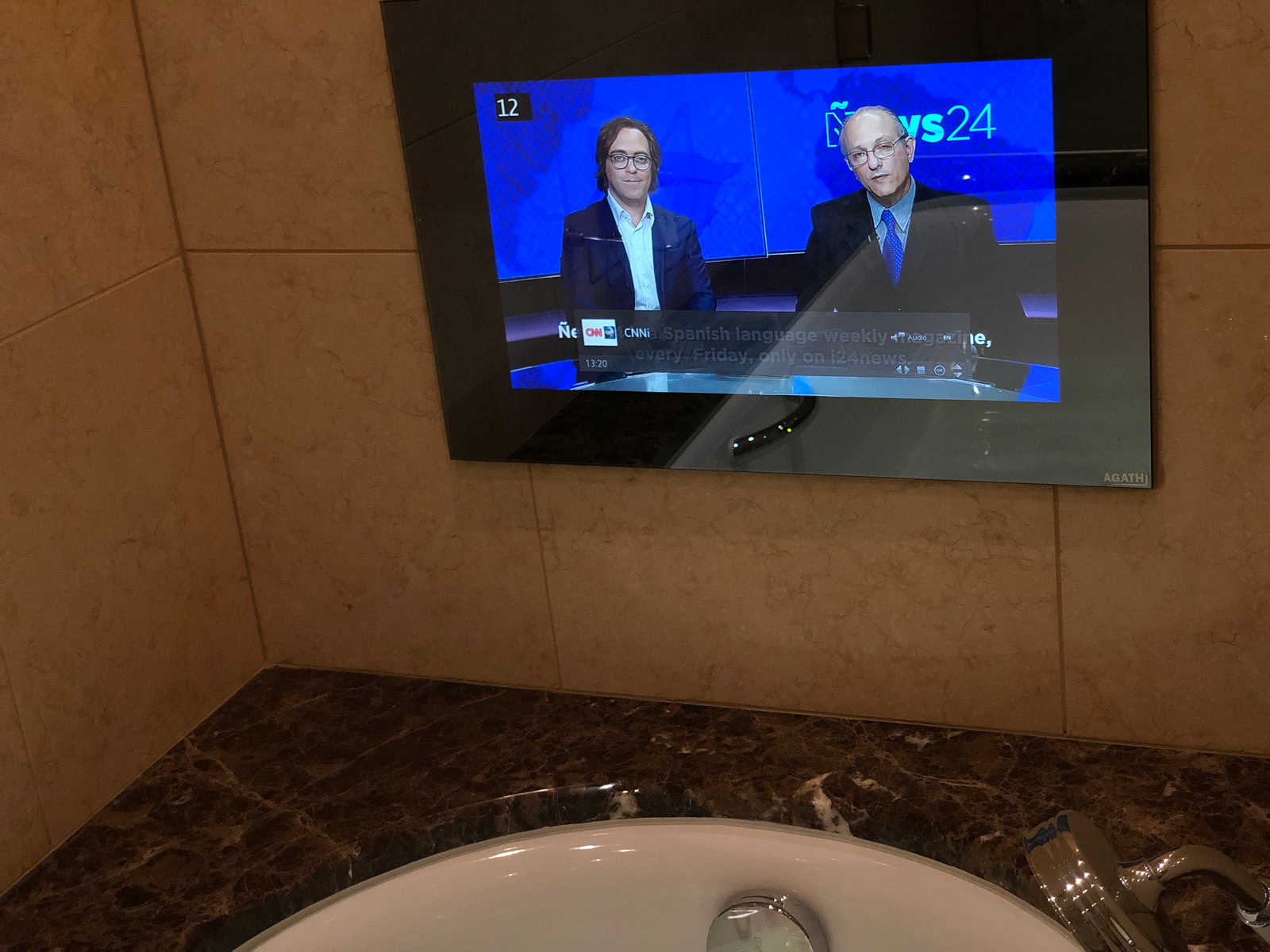 A bathroom mirror with an integrated TV displaying a news program, situated above a white sink and dark marble counter.