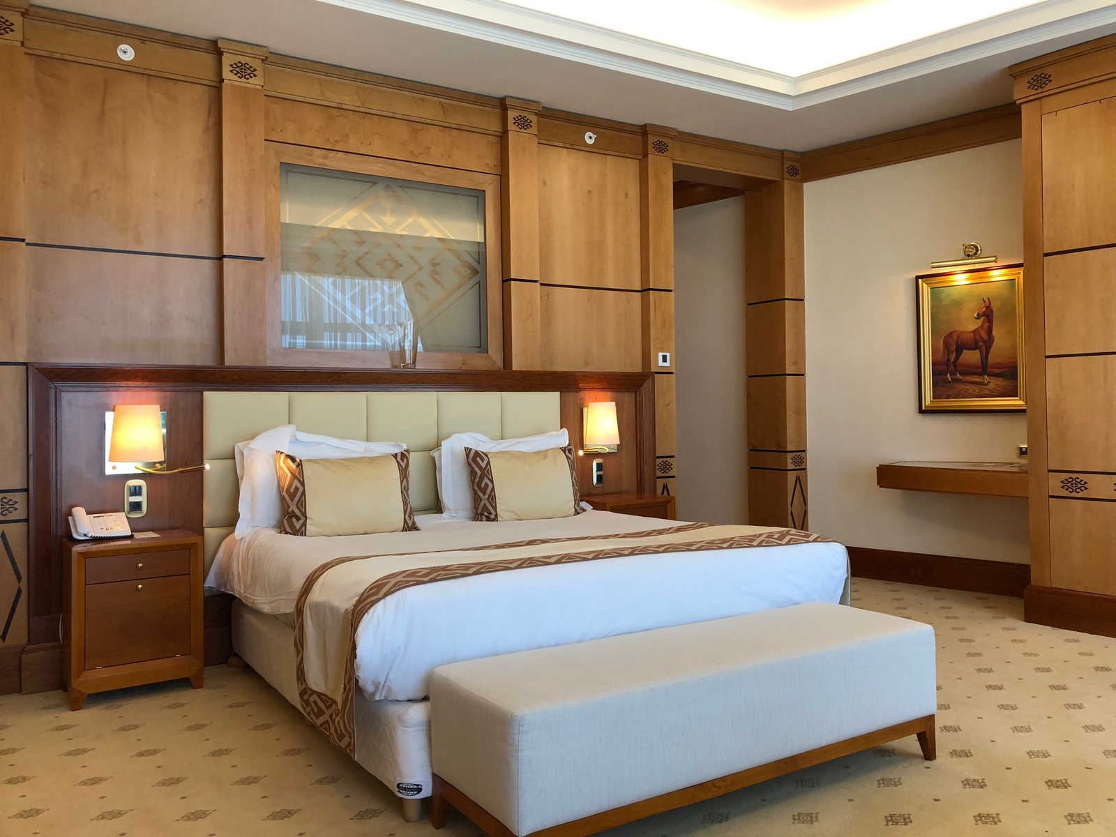 A luxurious hotel bedroom with a large bed, wooden paneling, a horse painting, and a bench at the foot of the bed.
