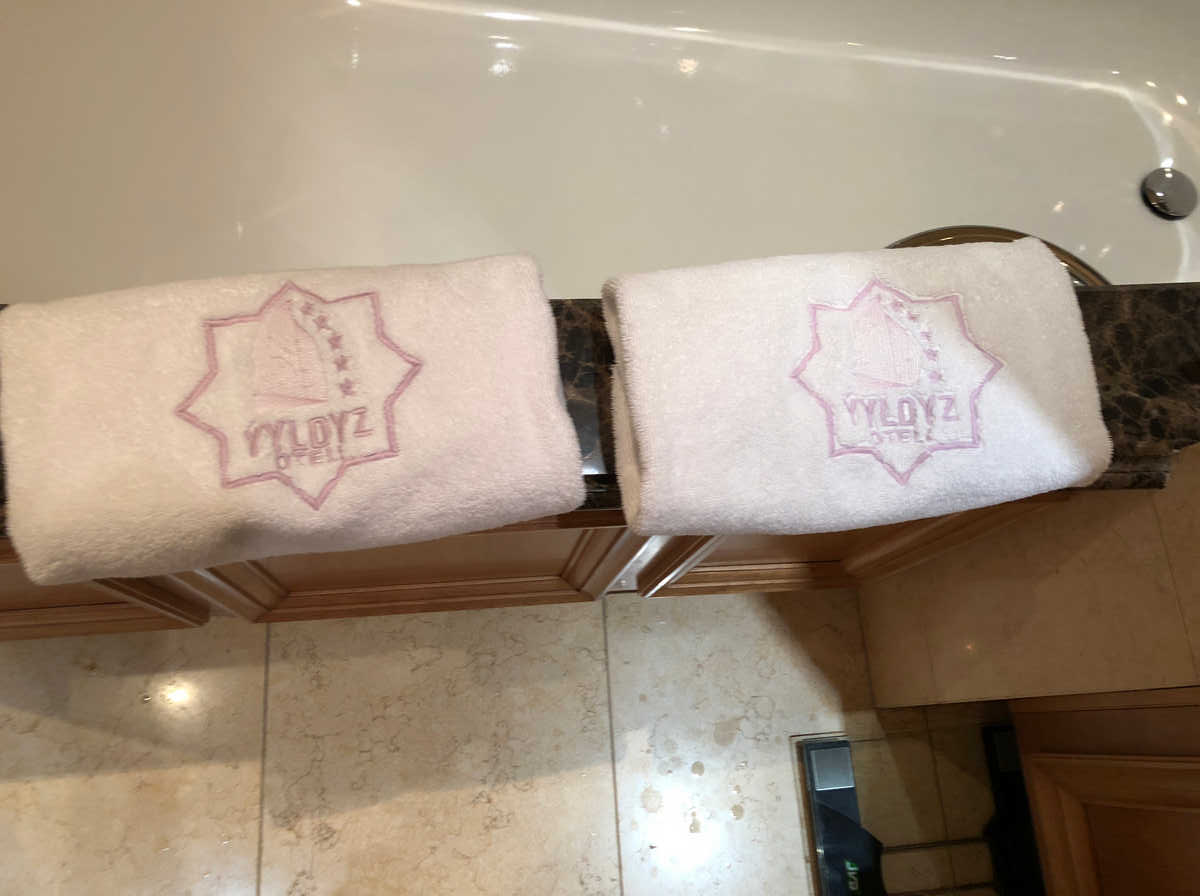 Two white towels with the pink Yyldyz Hotel logo are neatly folded on a dark countertop next to a white bathtub.