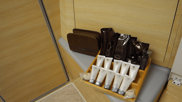 A brown amenities kit bag and various toiletries on a wooden shelf in an Emirates First Class cabin.