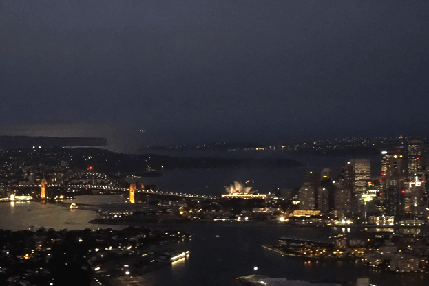 Aerial night view of Sydney's illuminated cityscape, including the Harbour Bridge and Opera House.