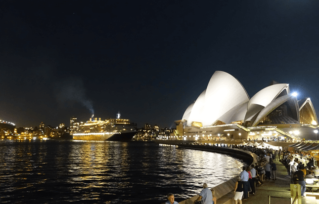 Night view of the illuminated Sydney Opera House and a cruise ship, with people gathered along the waterfront.