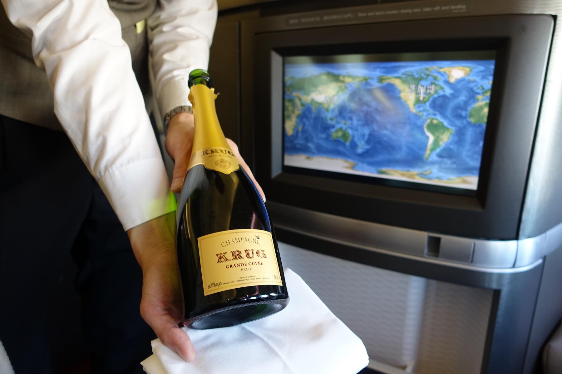A flight attendant presents a bottle of Krug champagne in front of an in-flight screen showing a world map.