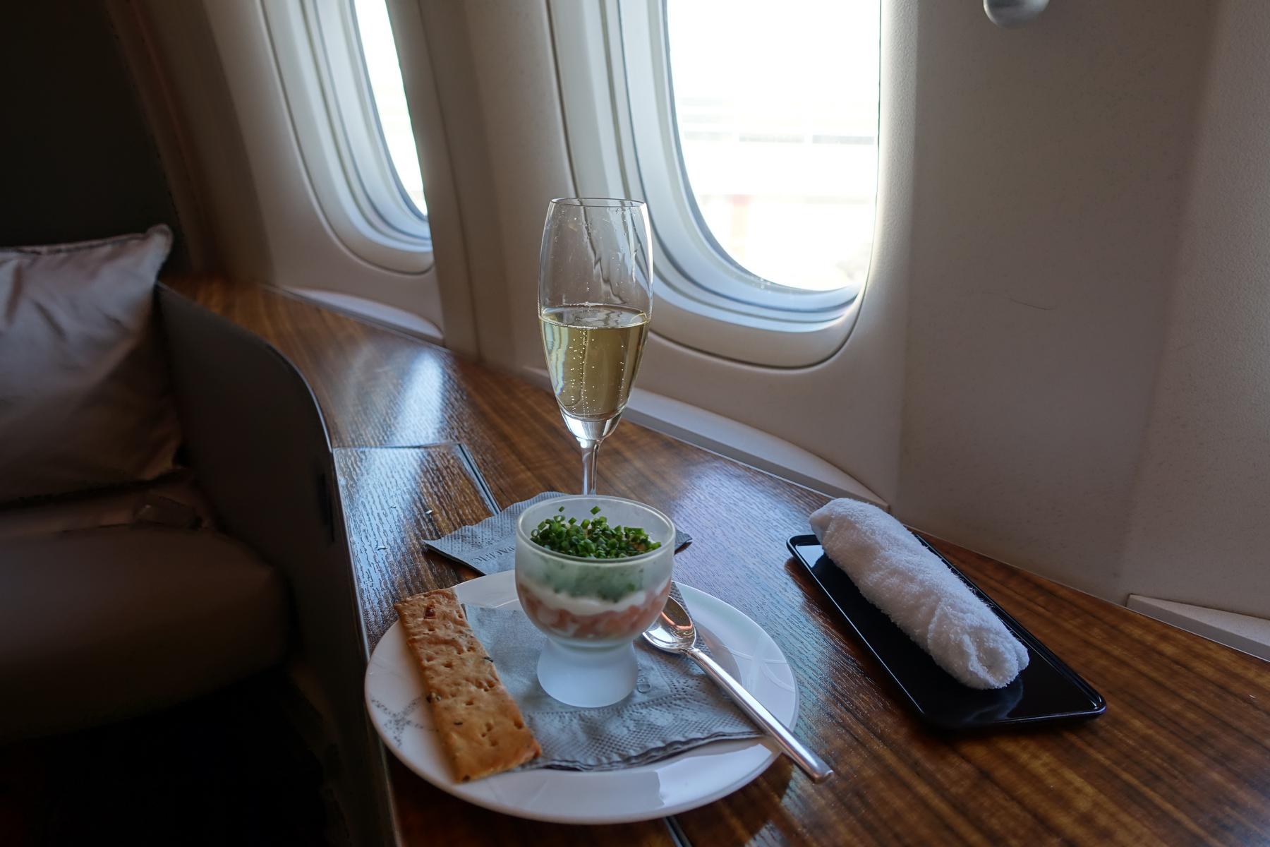 A flute of champagne and a layered seafood appetizer with crackers are served on a wooden table in an airplane's first-class cabin.