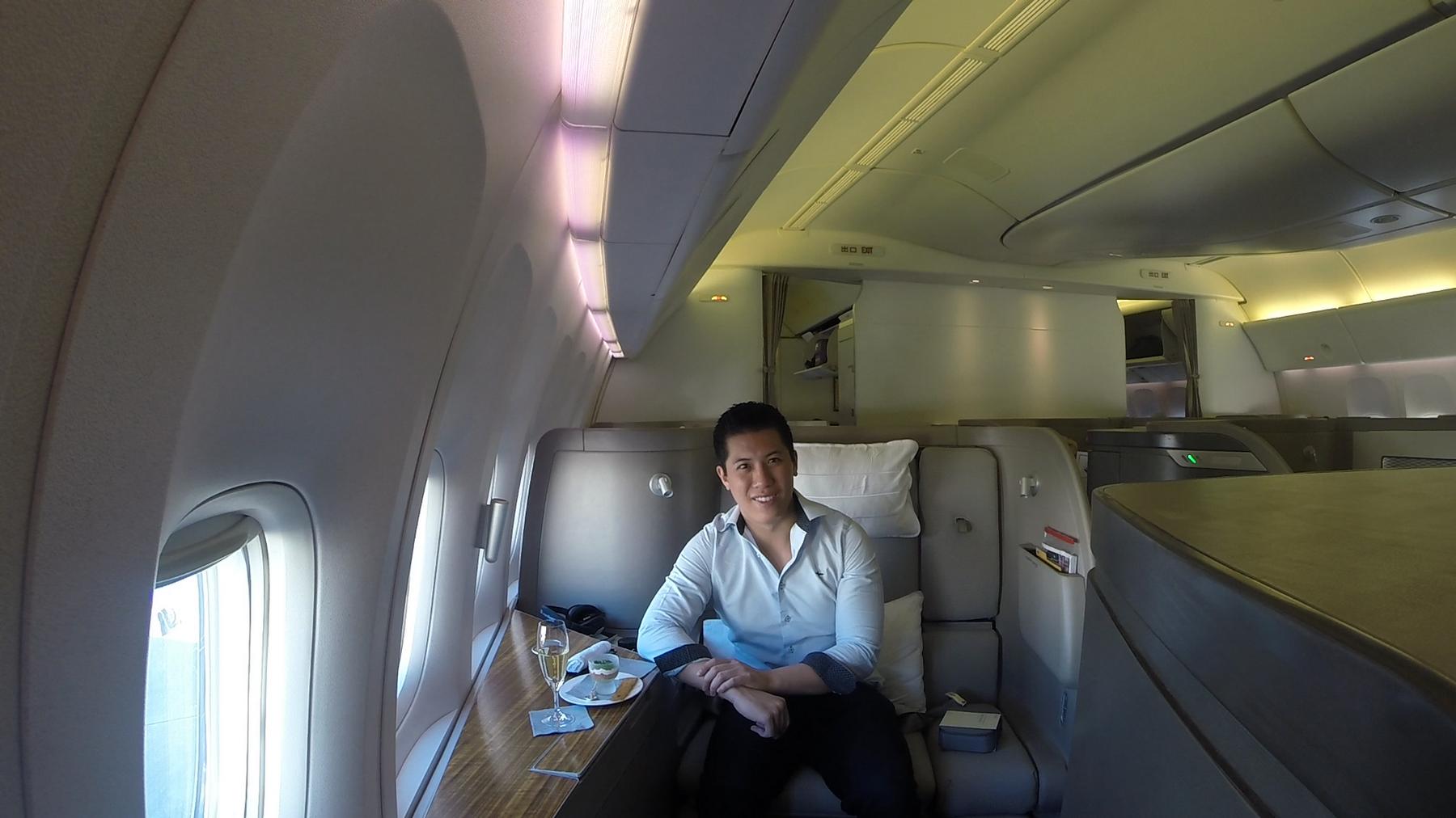 A smiling man sits in a spacious Cathay Pacific First Class seat next to a window, with champagne.