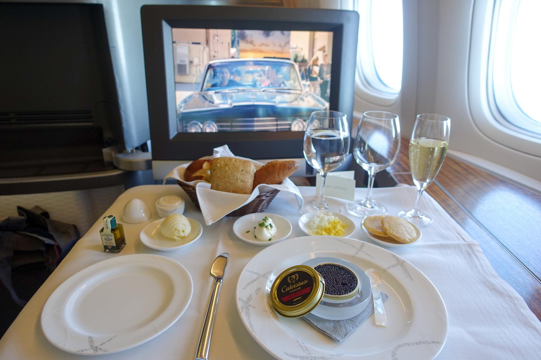 A lavish First Class airline meal featuring caviar, champagne, and bread, served on a white tablecloth with a movie playing on the personal screen.