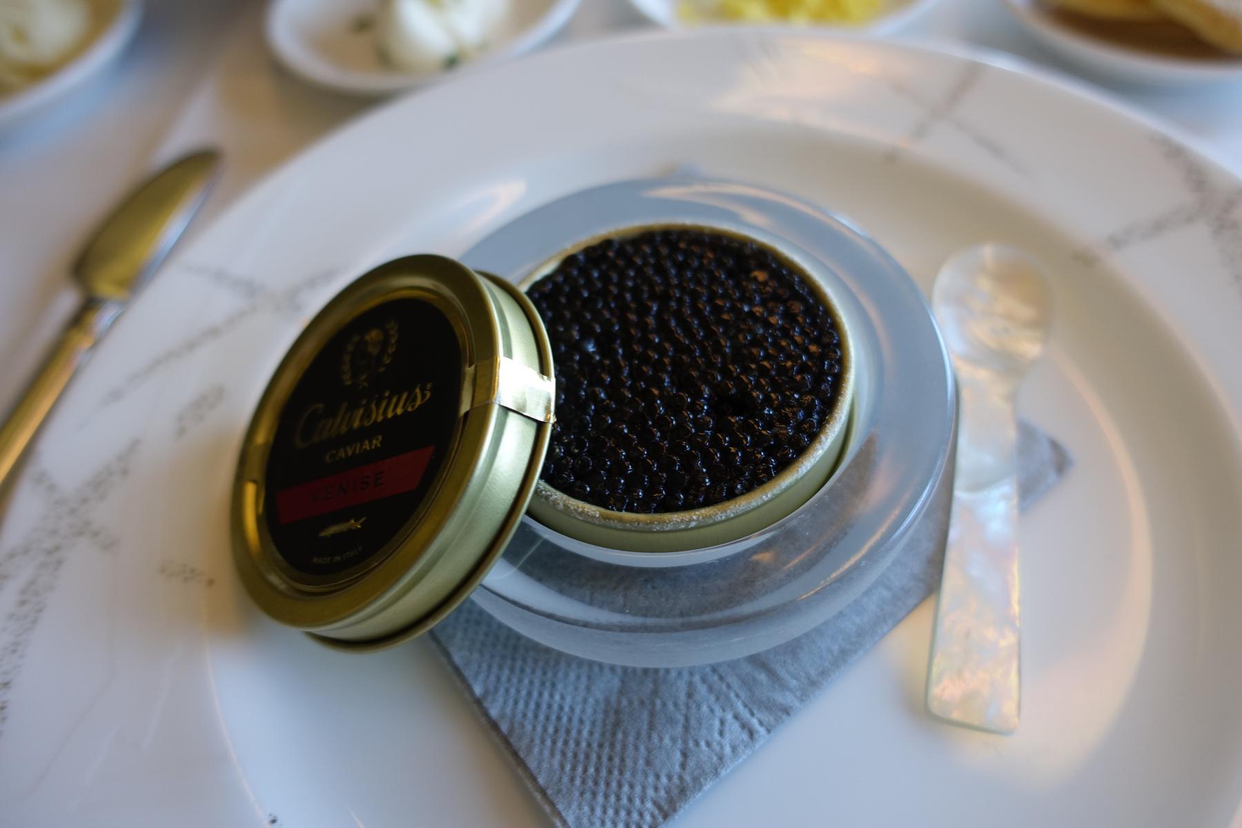 An open tin of black caviar is served with a mother-of-pearl spoon on a white plate.