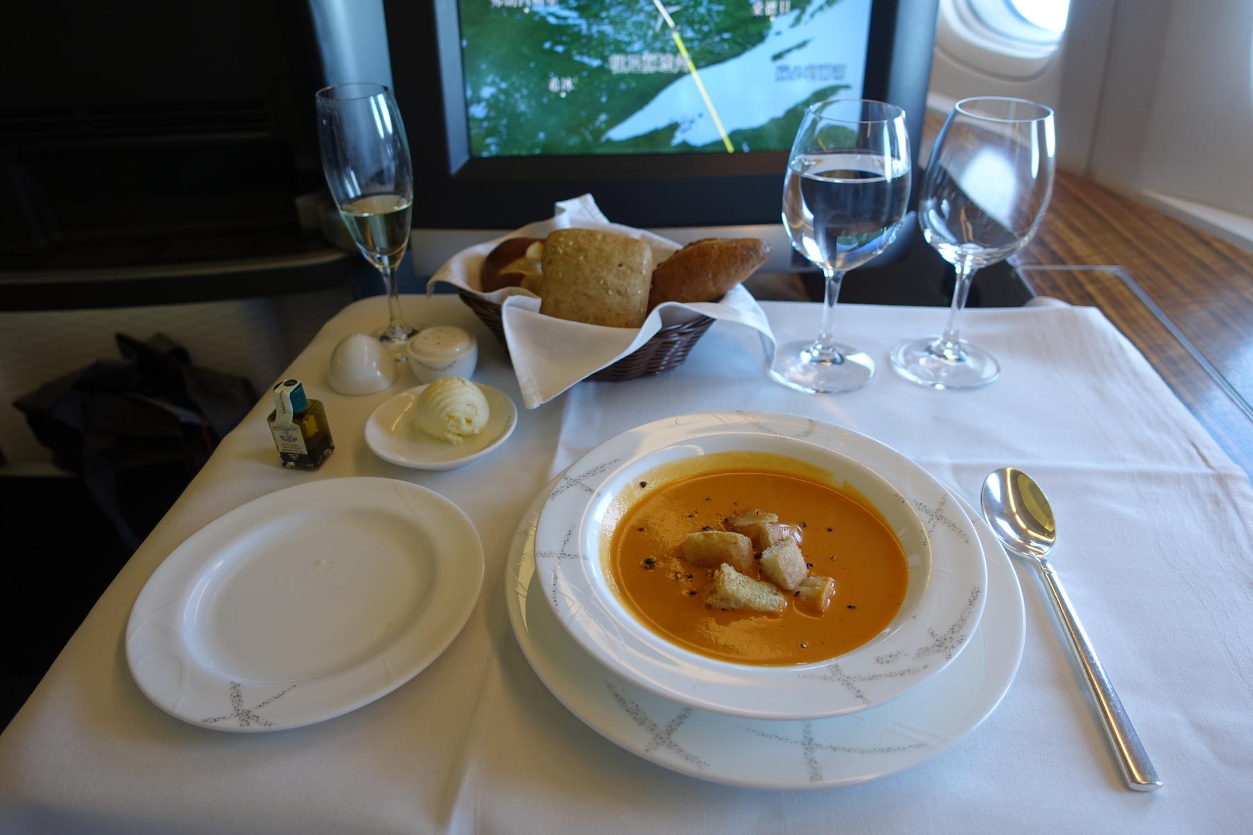 A first-class airplane meal is displayed on a table with a white tablecloth, featuring a bowl of orange soup with croutons, a bread basket, butter, champagne, and water glasses, with a flight map visible on a screen.