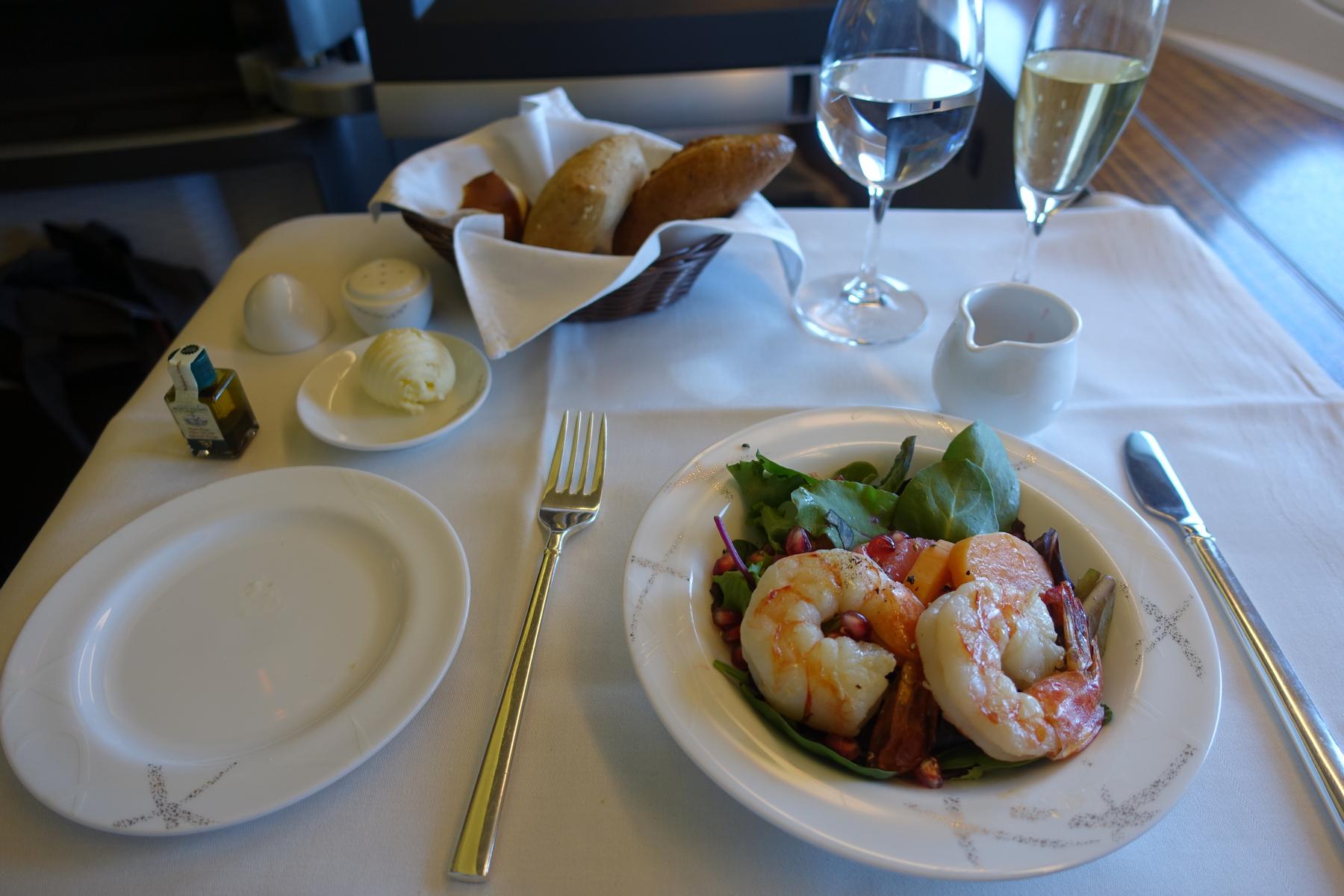 A first-class airplane meal with shrimp and salad, a bread basket, and two glasses of drinks served on a white tablecloth.