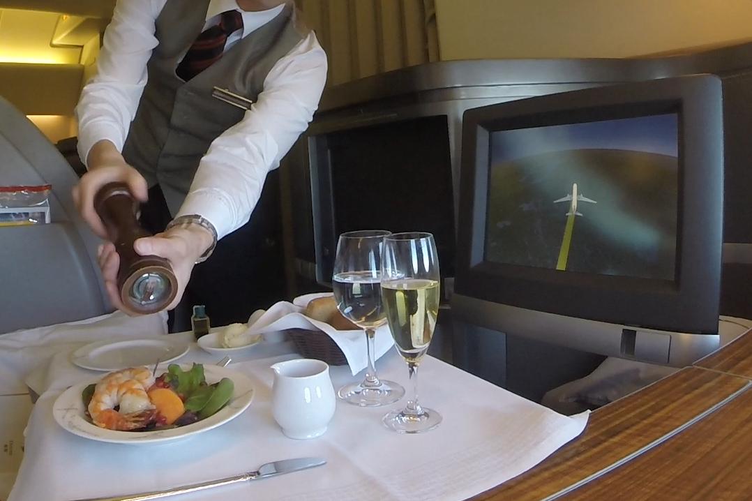 A flight attendant grinds pepper onto a shrimp and salad meal in Cathay Pacific First Class, with an airplane map on the screen.