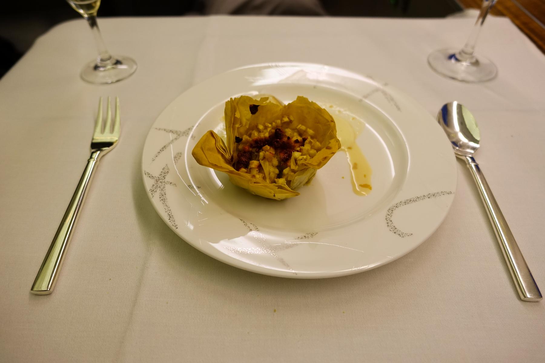 A gourmet dessert in a golden pastry cup on a white plate with cutlery, served on a white tablecloth, from a first-class flight.