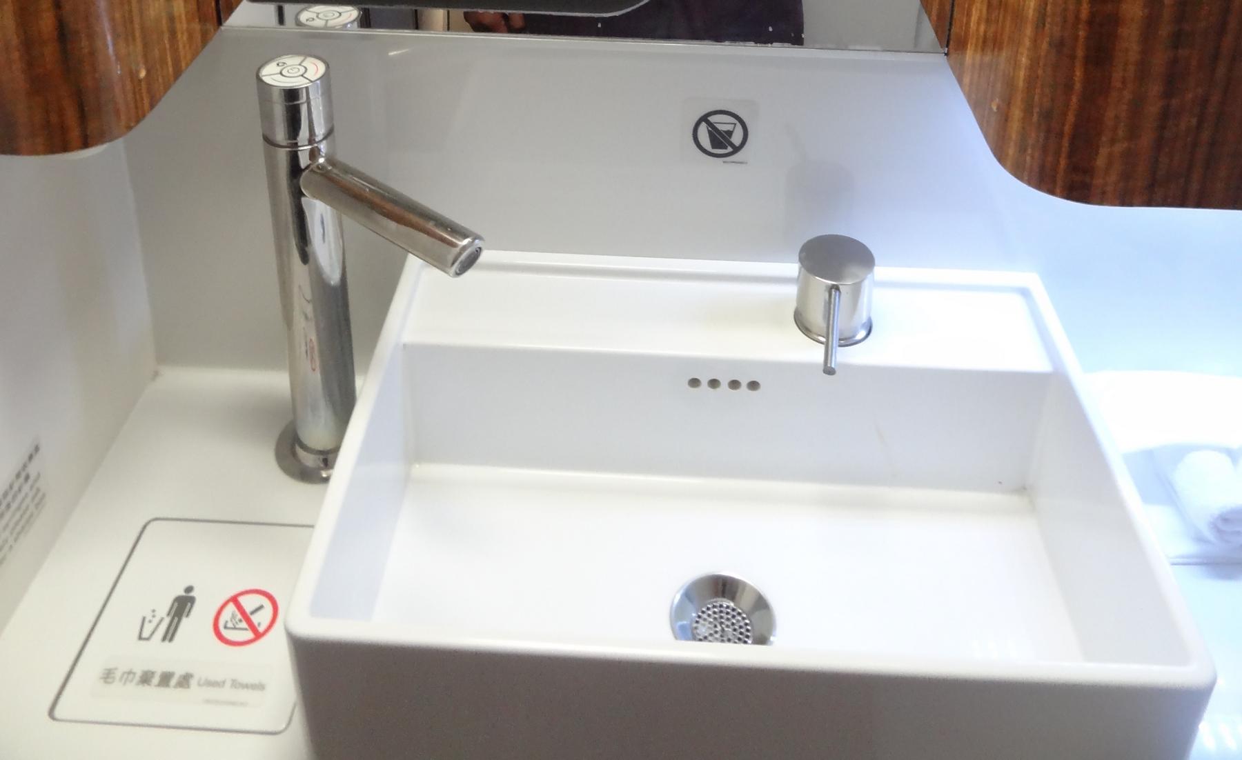 A sleek white rectangular sink with a tall chrome faucet and a chrome soap dispenser in a Cathay Pacific First Class lavatory.