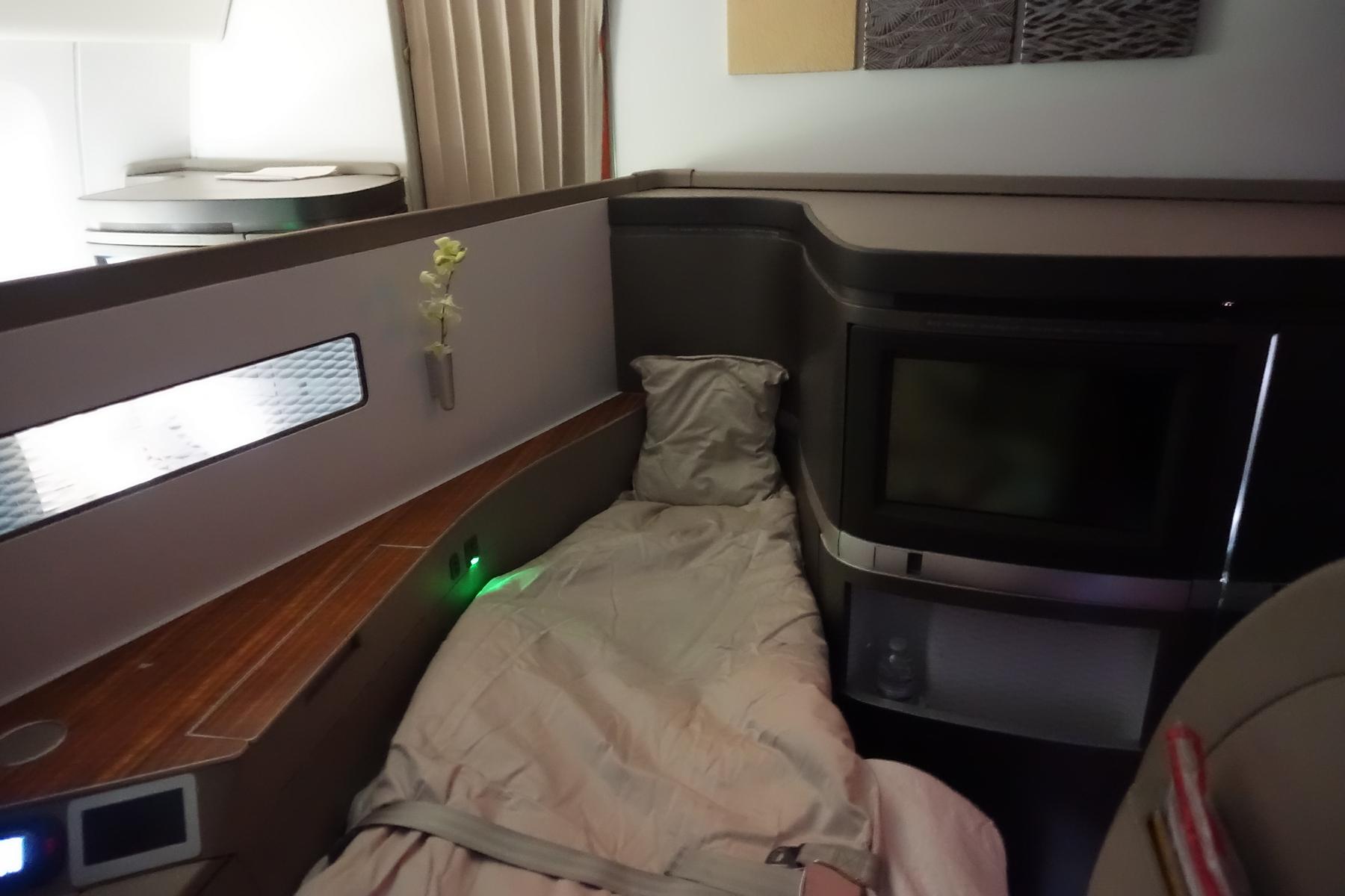 An empty Cathay Pacific First Class suite features a lie-flat bed, TV screen, and small white flowers.