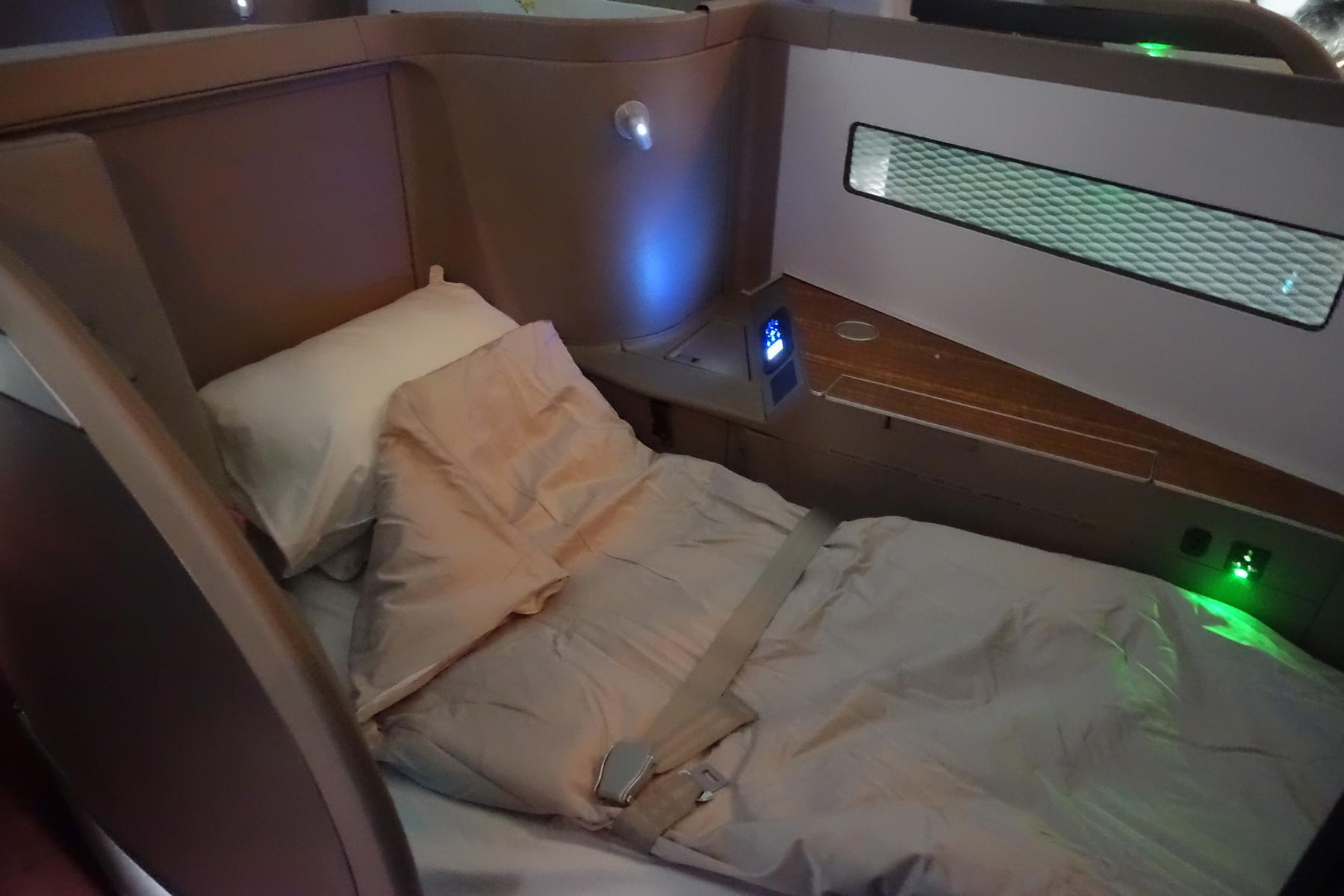 A Cathay Pacific First Class seat fully reclined into a comfortable bed with a pillow, duvet, and seatbelt.