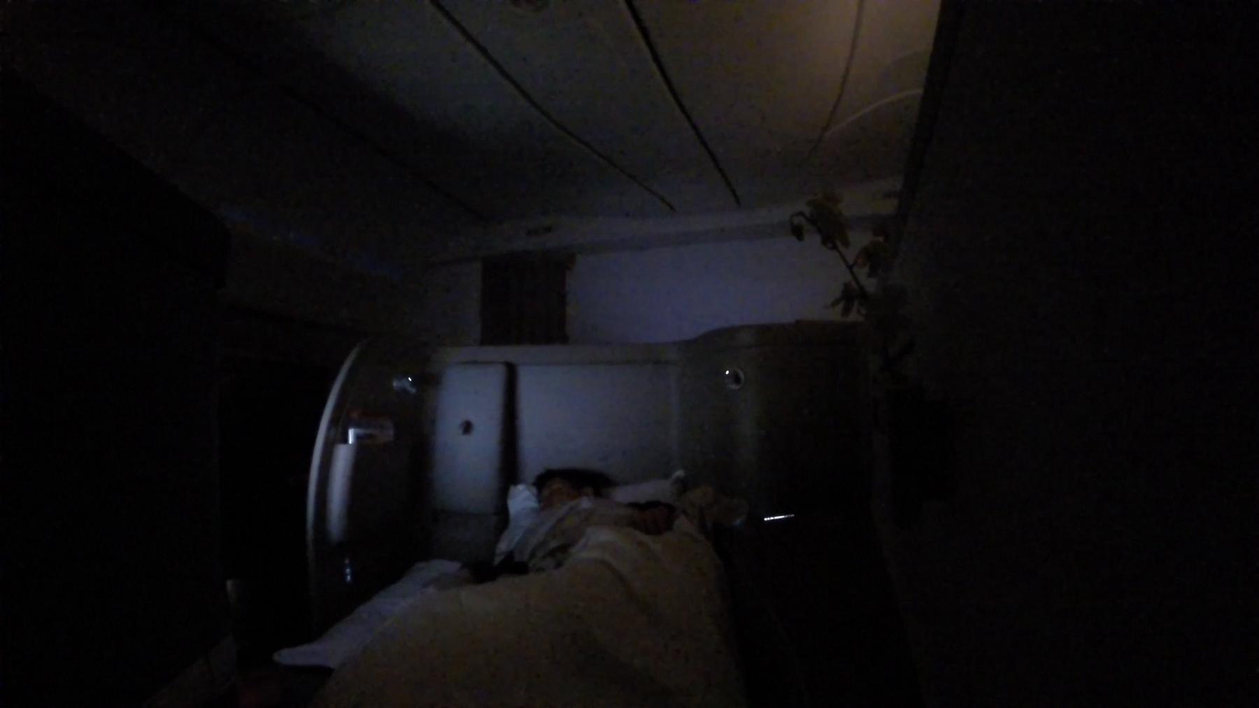 A passenger sleeps in a flat bed in a dimly lit first class airplane cabin.