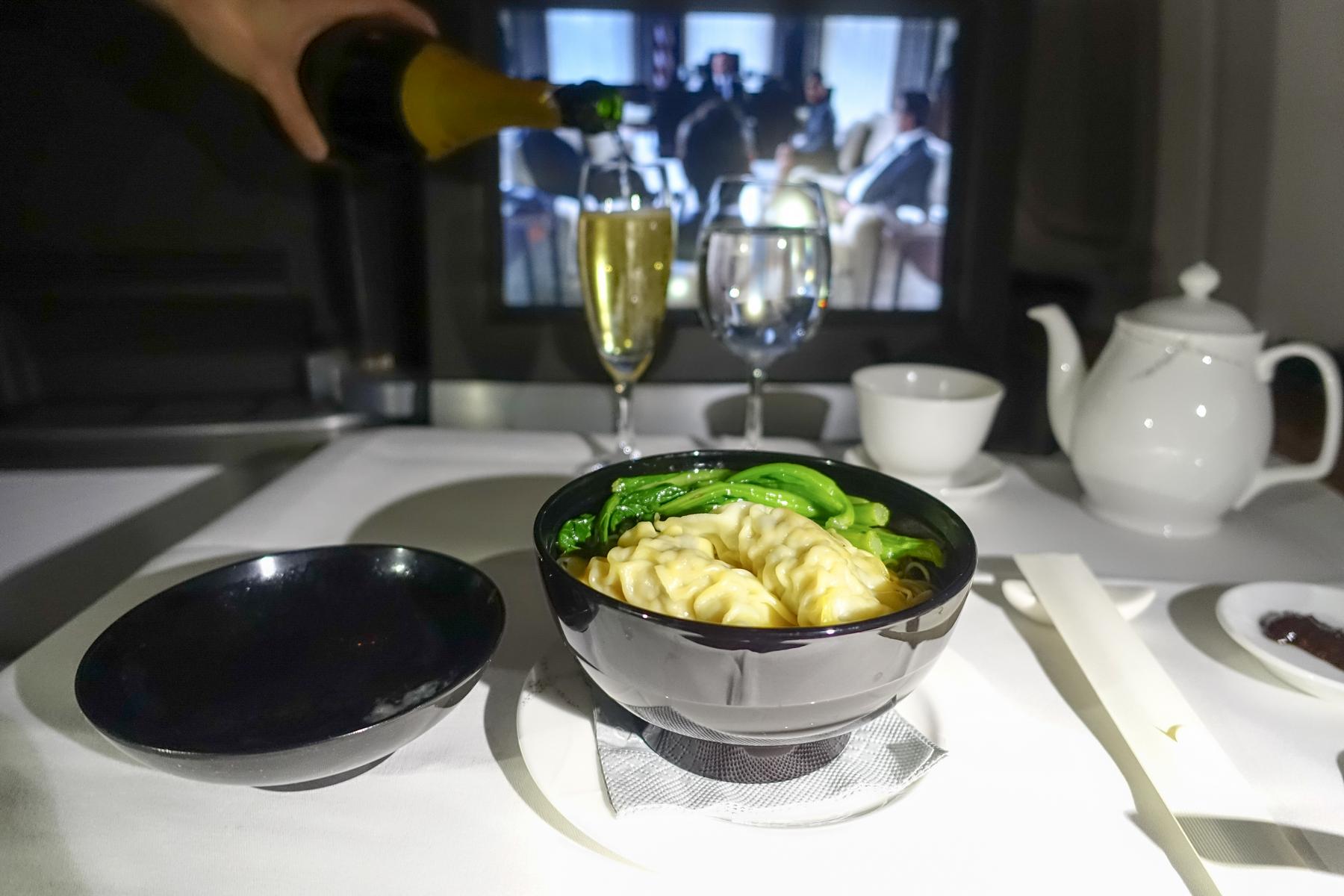 A hand pours champagne into a glass next to a bowl of wonton noodles and greens on an airplane tray table.
