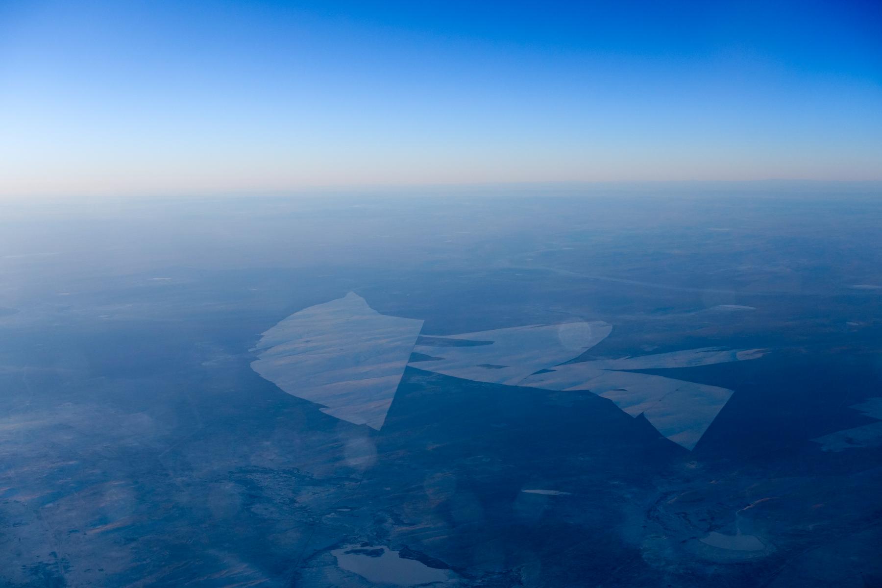 Aerial view of a hazy, blue-toned landscape below a clear sky, with prominent reflections on the airplane window.