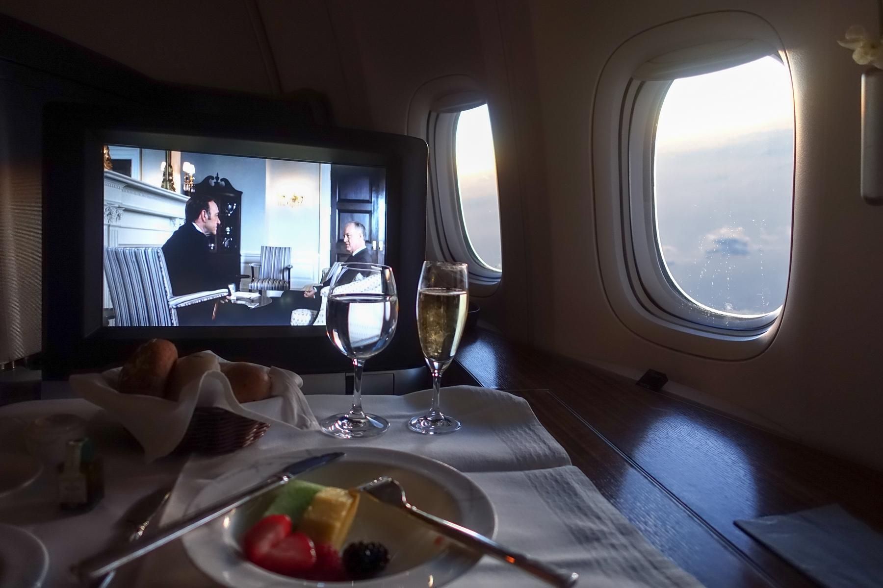 A first-class airplane cabin with a set table, drinks, a movie playing on screen, and a bright window view of clouds.