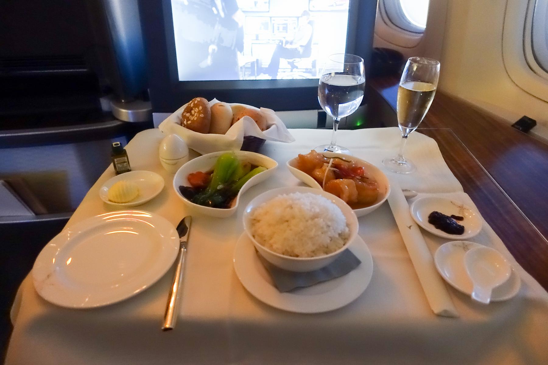 A luxurious multi-course meal with rice, vegetables, and champagne served on a white tablecloth in an airplane first class cabin.