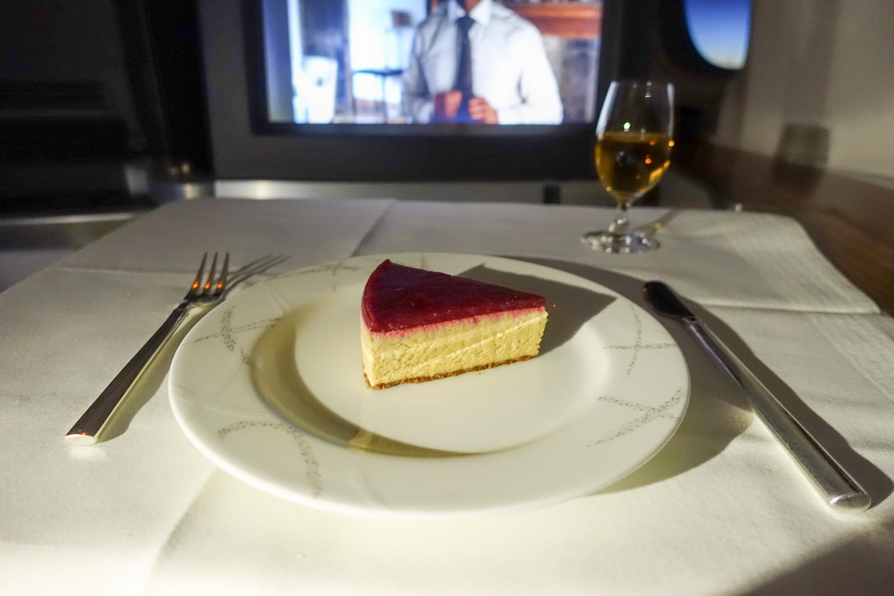 A slice of cheesecake with a red topping sits on a white plate with cutlery, a drink, and an in-flight entertainment screen in a First Class airplane cabin.