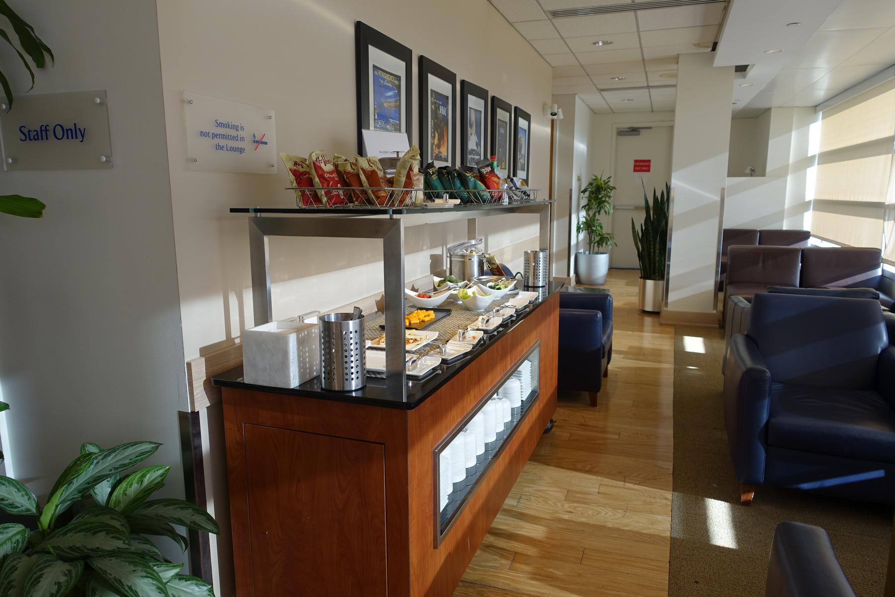 A well-lit airport lounge features a self-service buffet with various snacks and small dishes, alongside comfortable seating areas.
