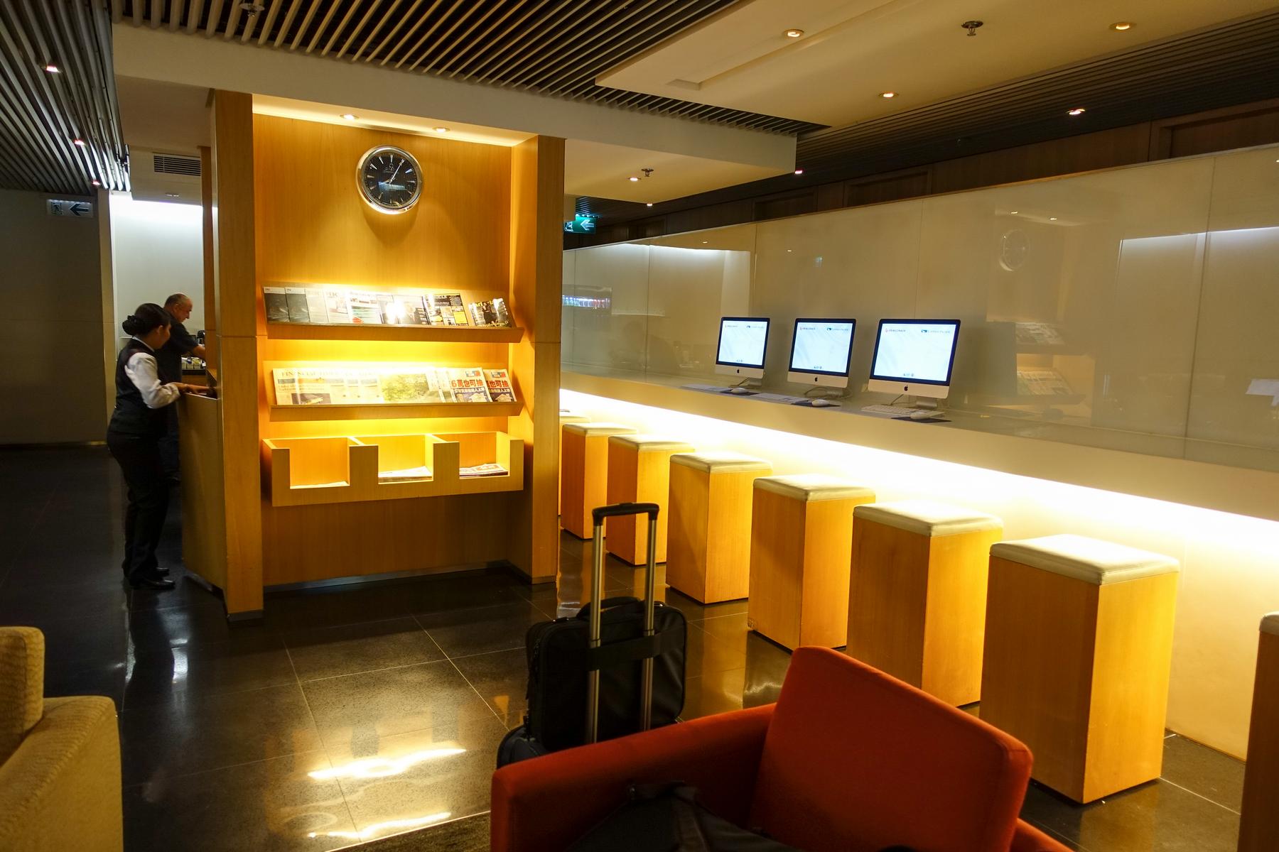 An airport lounge features a wooden magazine rack with a clock, a long counter with computer workstations, and a staff member at a desk.