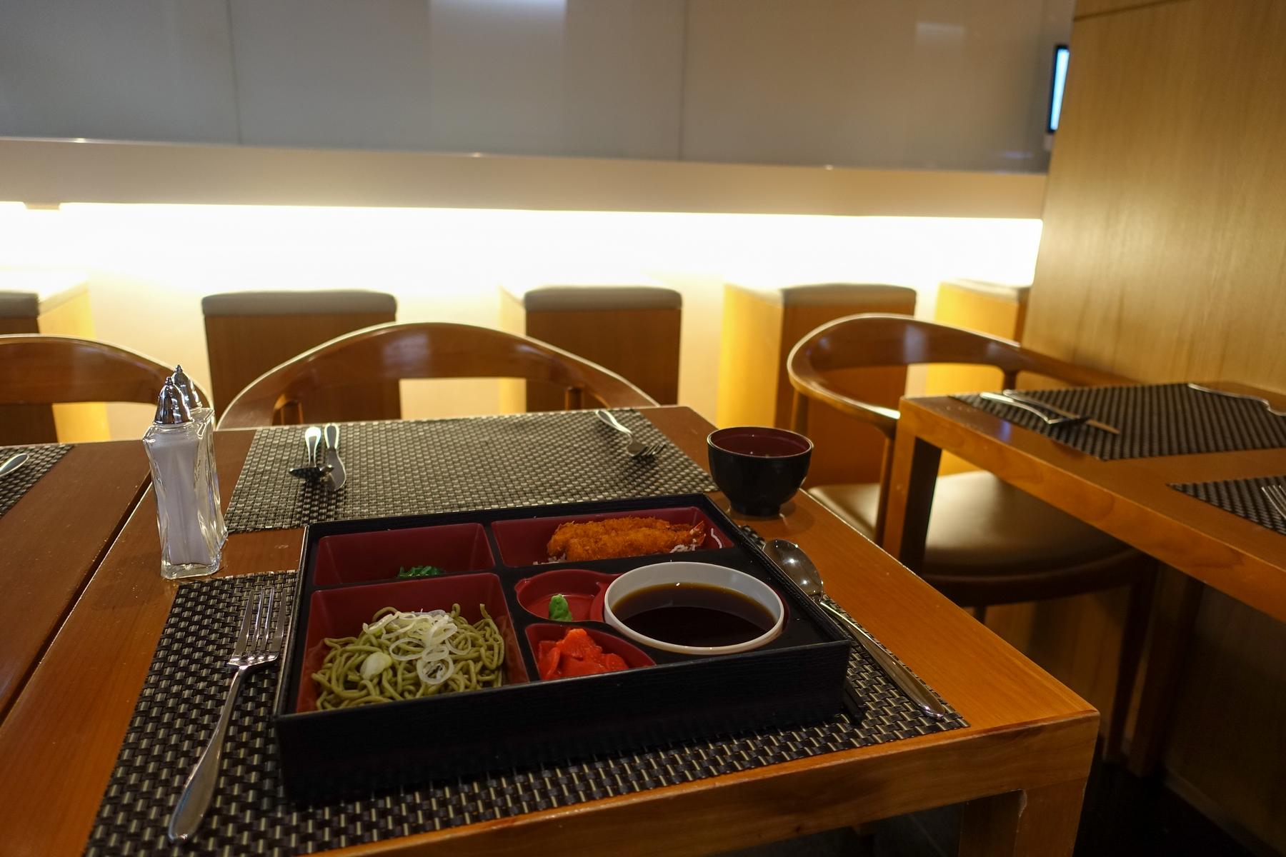 A Japanese bento box meal with green noodles, fried shrimp, and dipping sauce on a wooden table in a lounge setting.