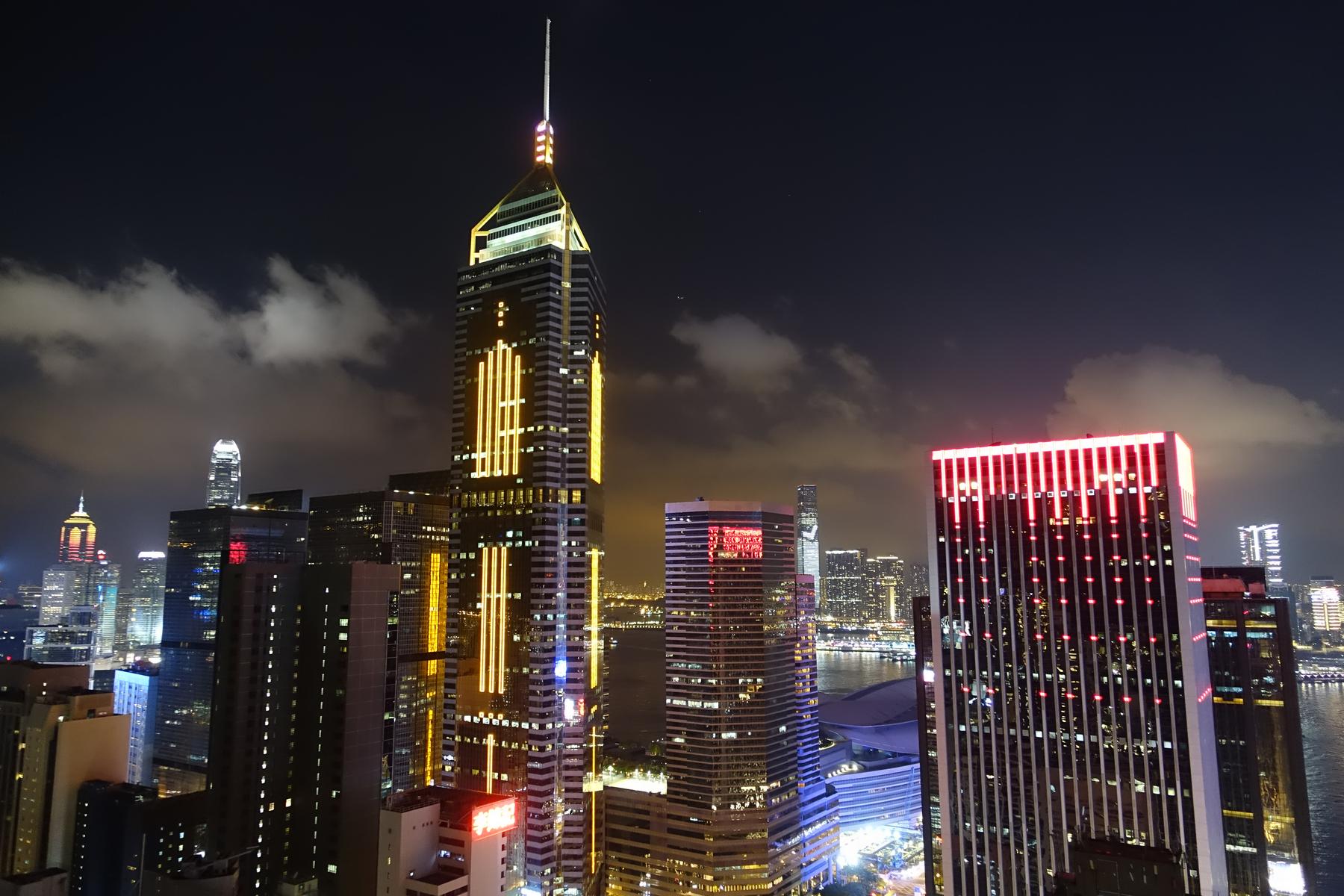 Nighttime view of the Hong Kong skyline with brightly lit skyscrapers and a visible harbor.