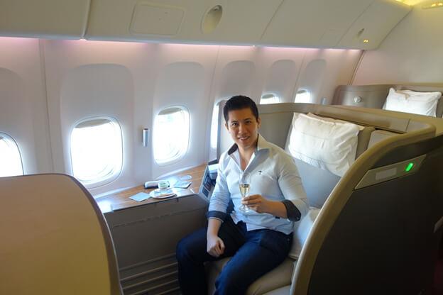 A man smiles while sitting in a spacious Cathay Pacific first-class airplane seat, holding a glass of champagne.