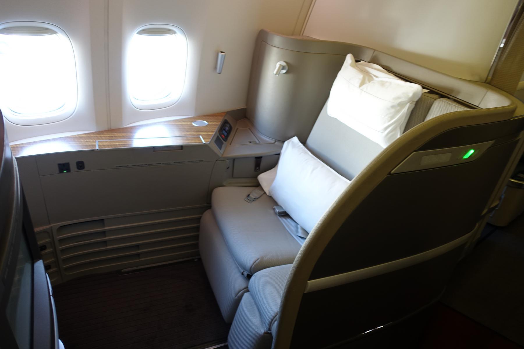 A spacious Cathay Pacific First Class airplane seat featuring two windows, a large seat with pillows, and a wooden console.