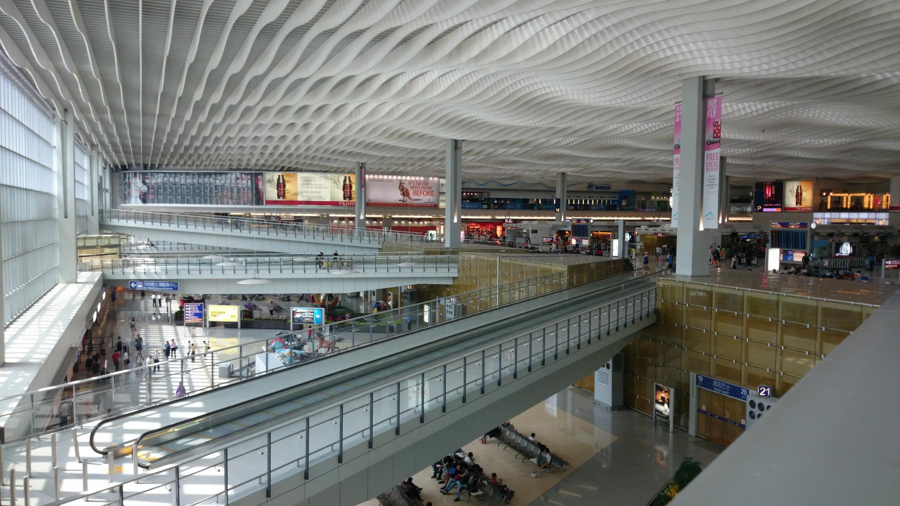 A large, multi-level airport terminal with a wavy white ceiling and long moving walkways.