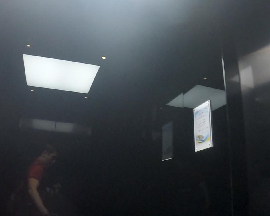 Dimly lit interior with a bright rectangular ceiling light, reflecting a person and a sign.
