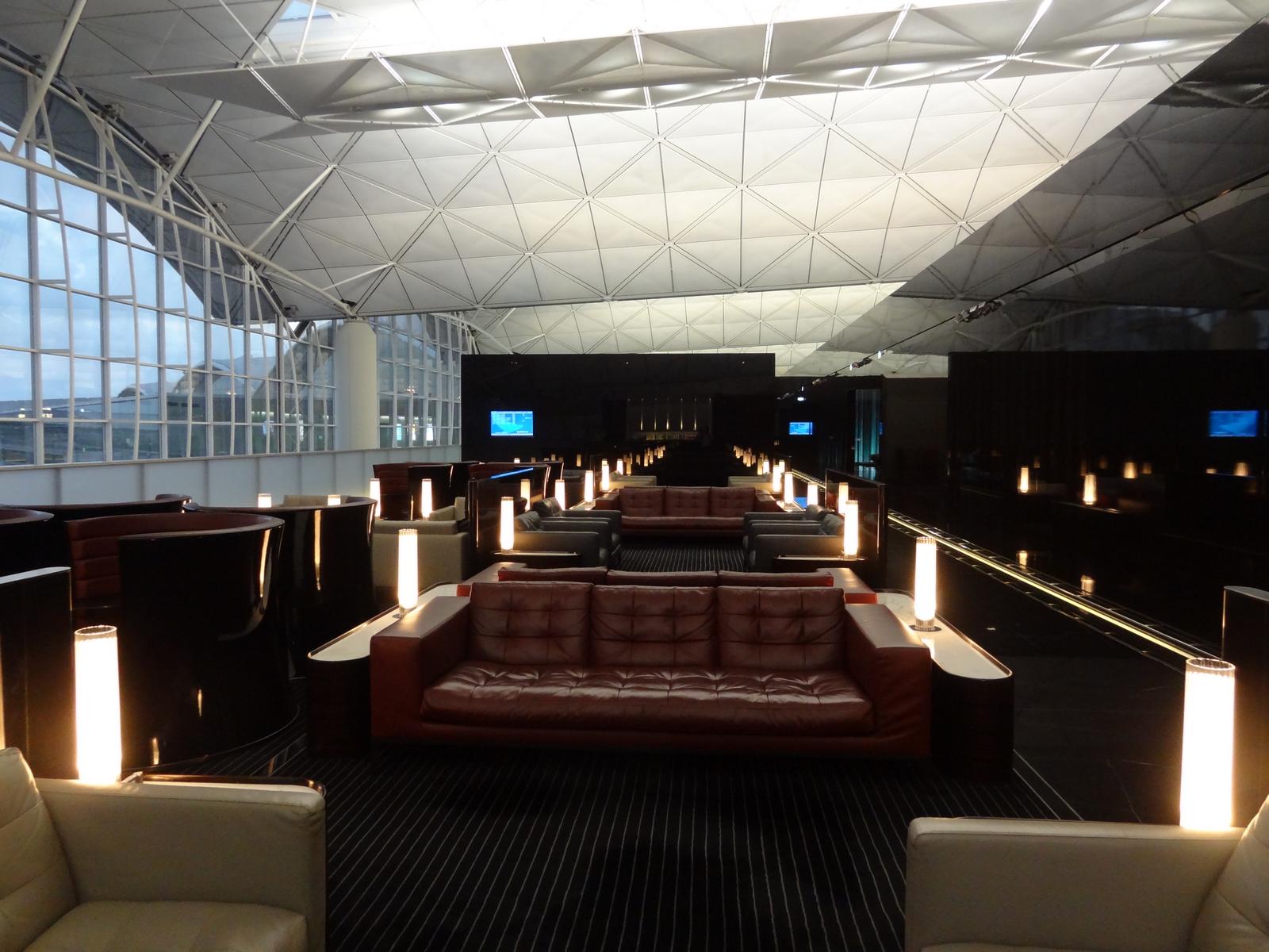 A modern Cathay First Class lounge with rows of leather sofas, bright pillar lamps, large windows, and a geometric ceiling.