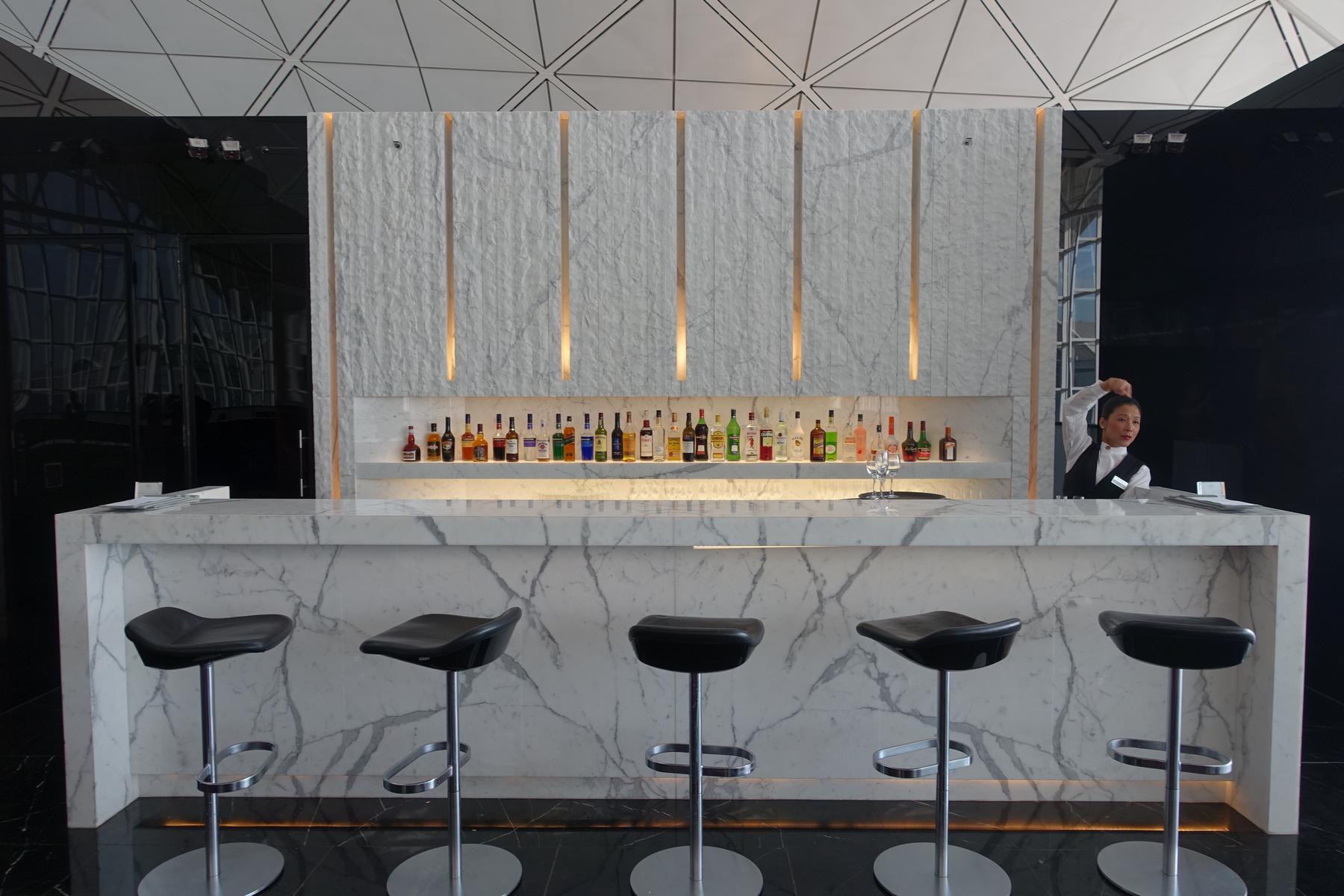 A white marble bar with a row of liquor bottles, five black stools, and a bartender.