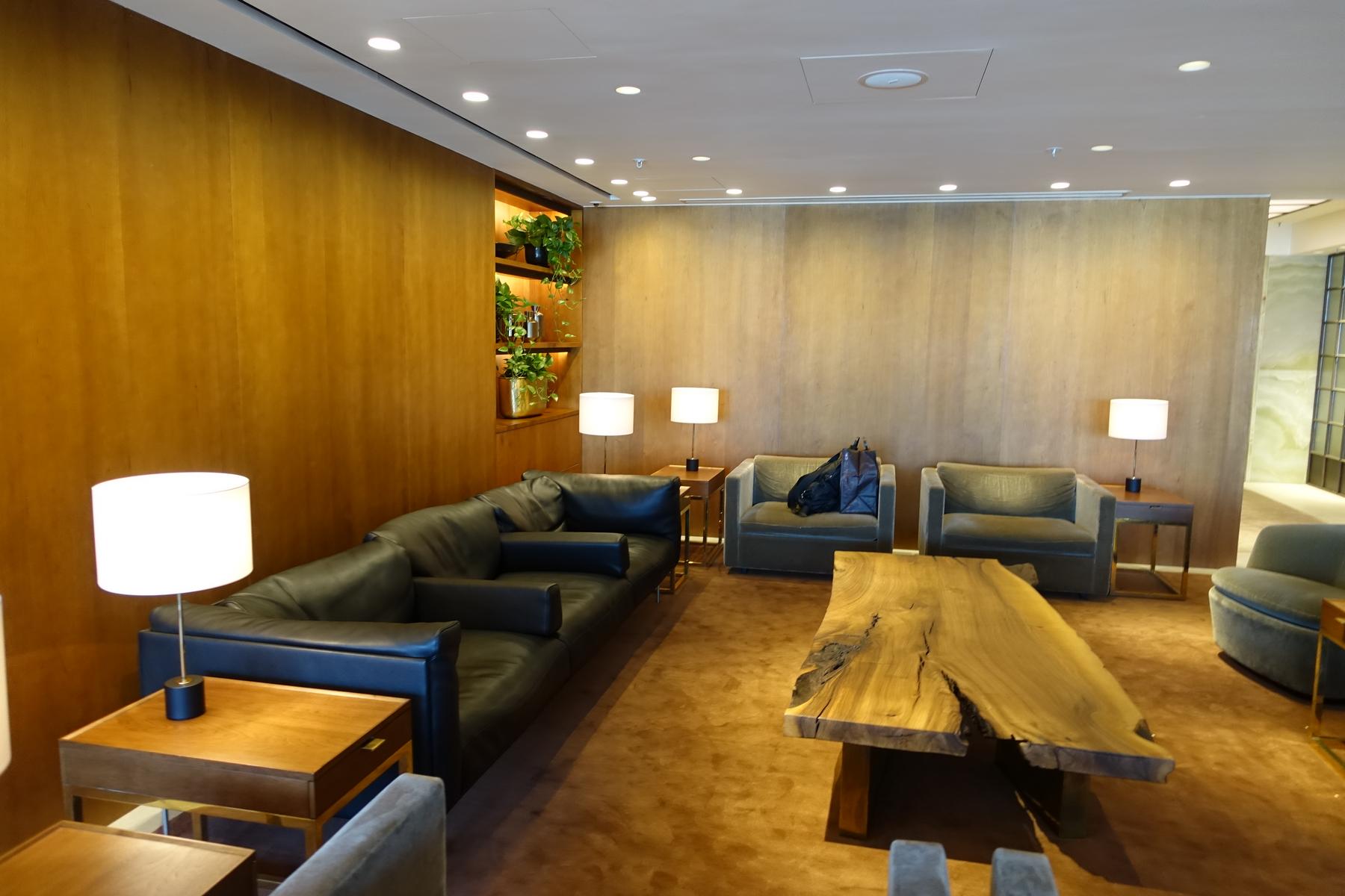 Cathay First Class Lounge seating area with dark leather sofas, armchairs, a large live-edge wooden coffee table, and wood-paneled walls.