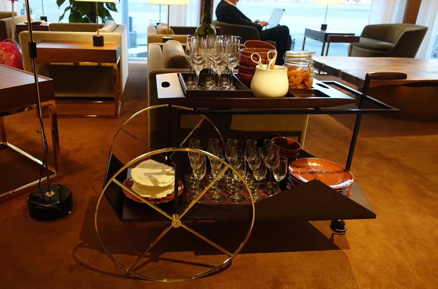 A stylish bar cart laden with wine, glasses, and snacks sits in a luxurious airport lounge.