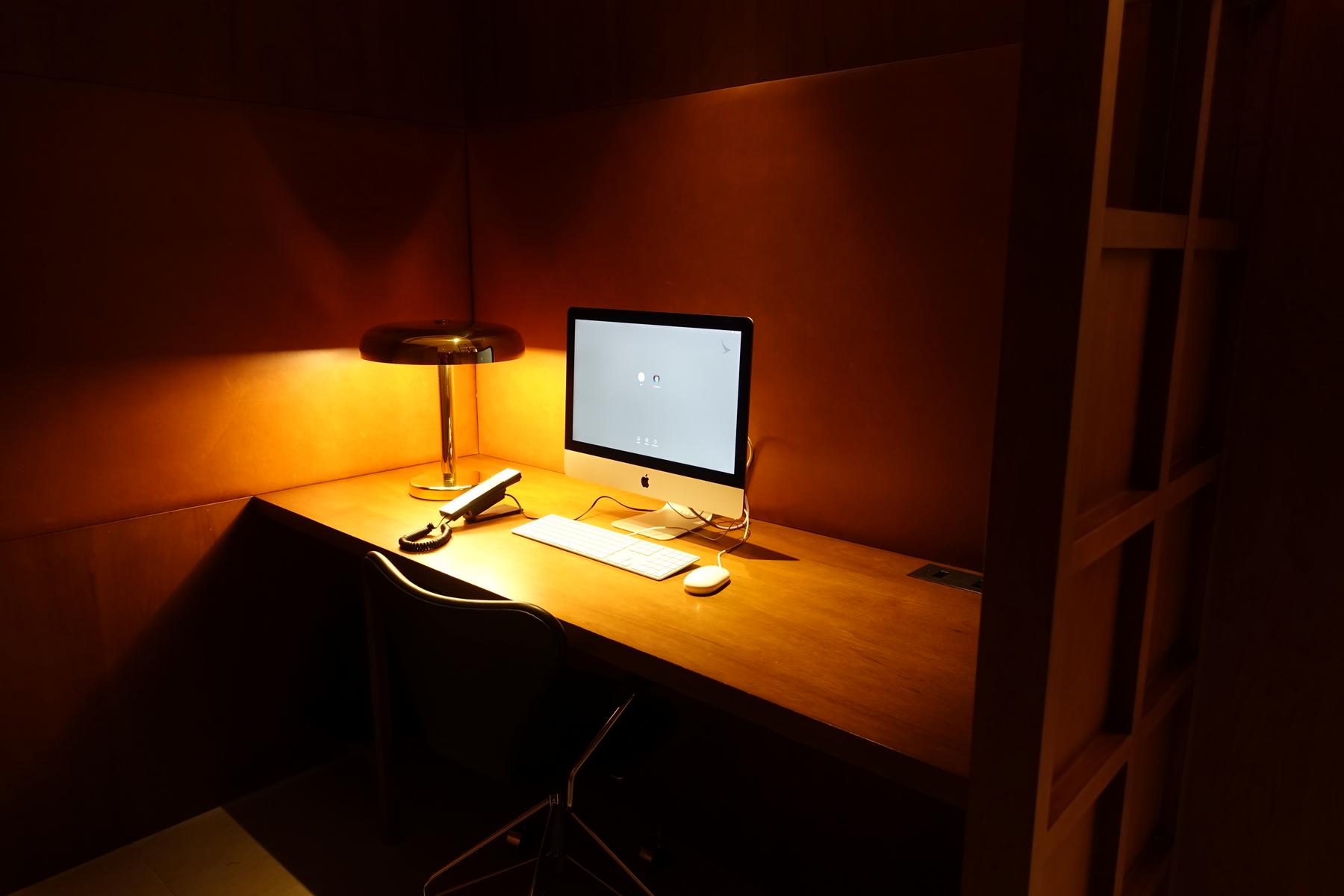 A dimly lit private cubicle with a desk, iMac, lamp, and phone in a first class lounge.