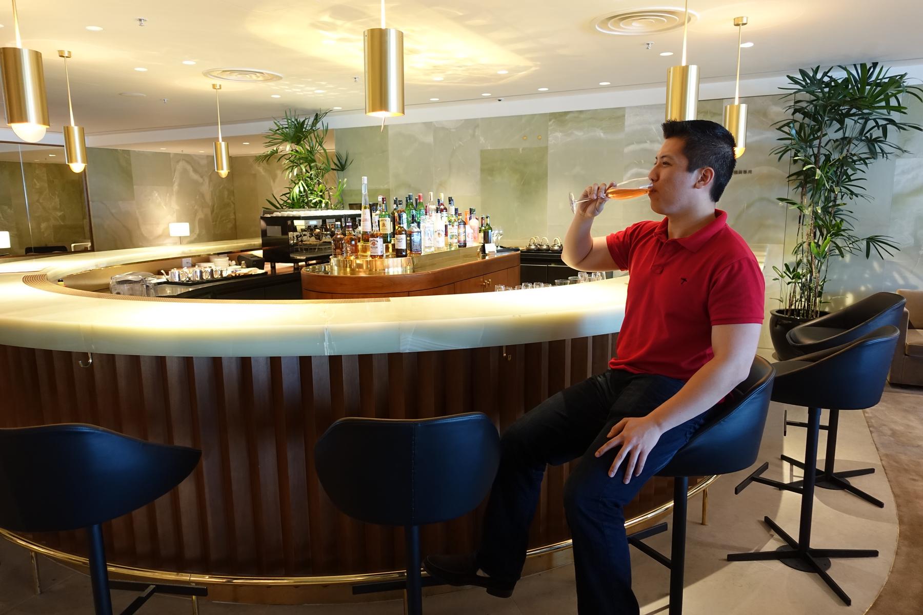 A man in a red polo shirt sips a drink at a curved bar in the Cathay First Class Lounge.