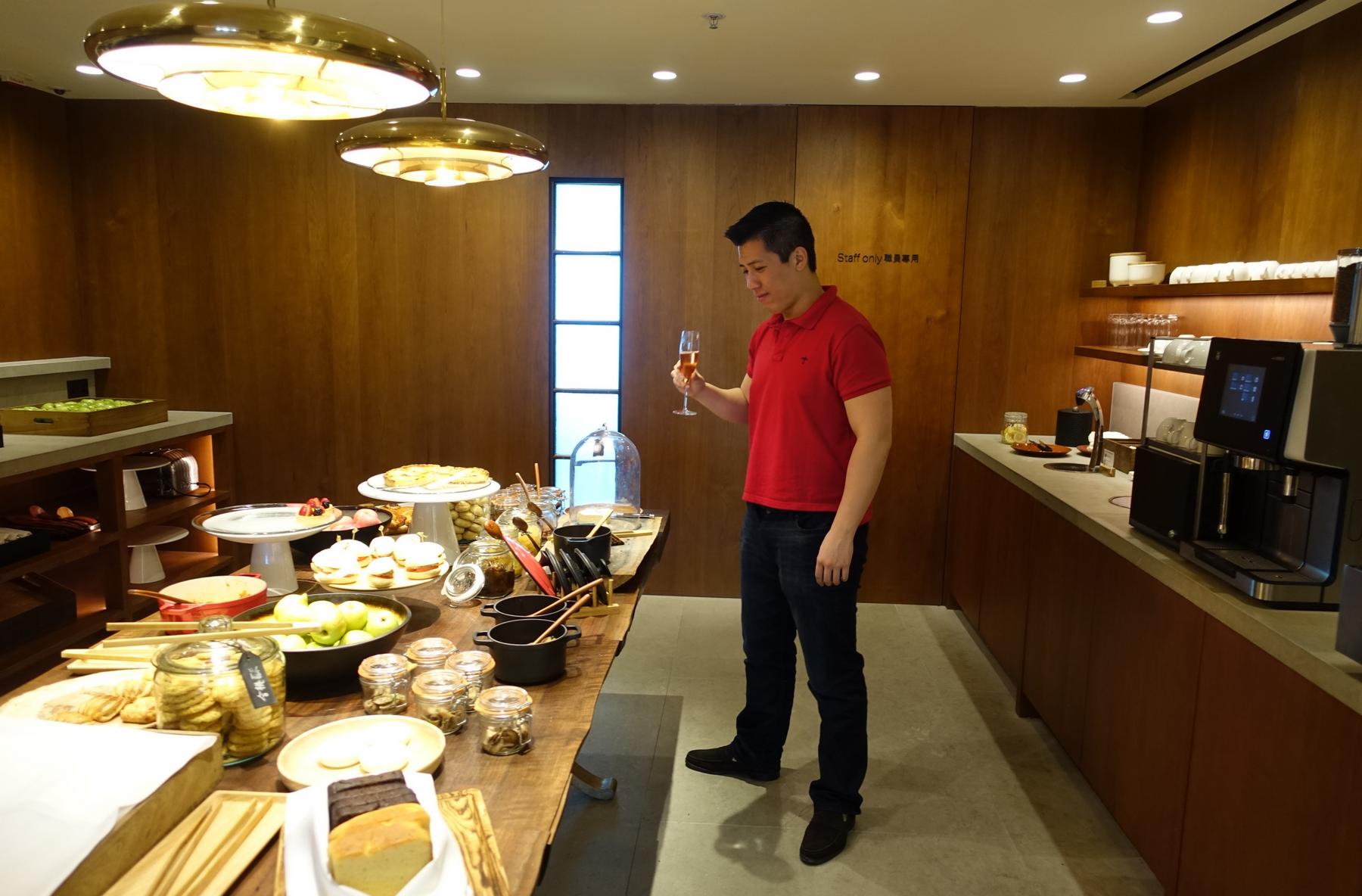 A man in a red polo shirt holds a glass of champagne next to a buffet table filled with pastries and snacks in a modern lounge.