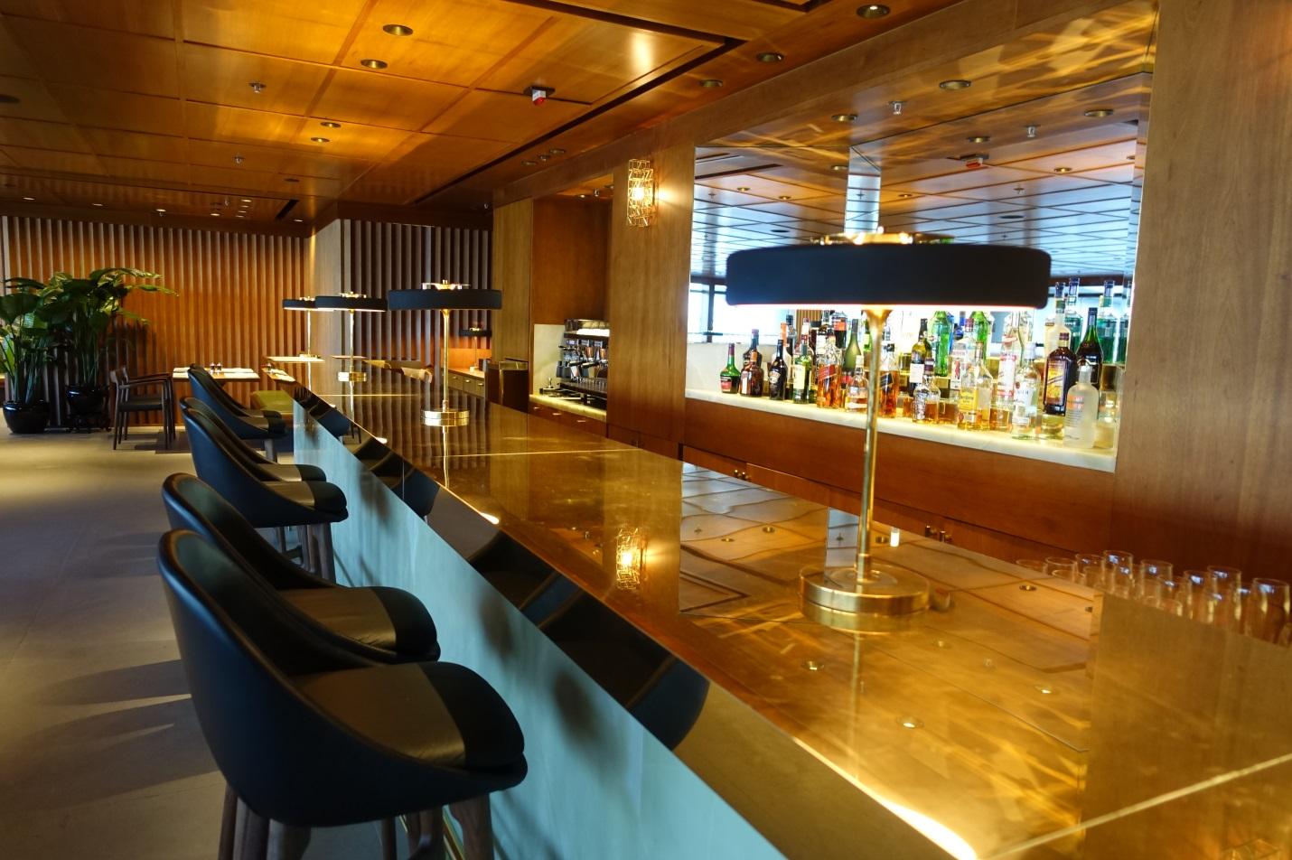 A long, gleaming golden bar counter with dark stools and a display of liquor bottles in a warm-toned lounge.