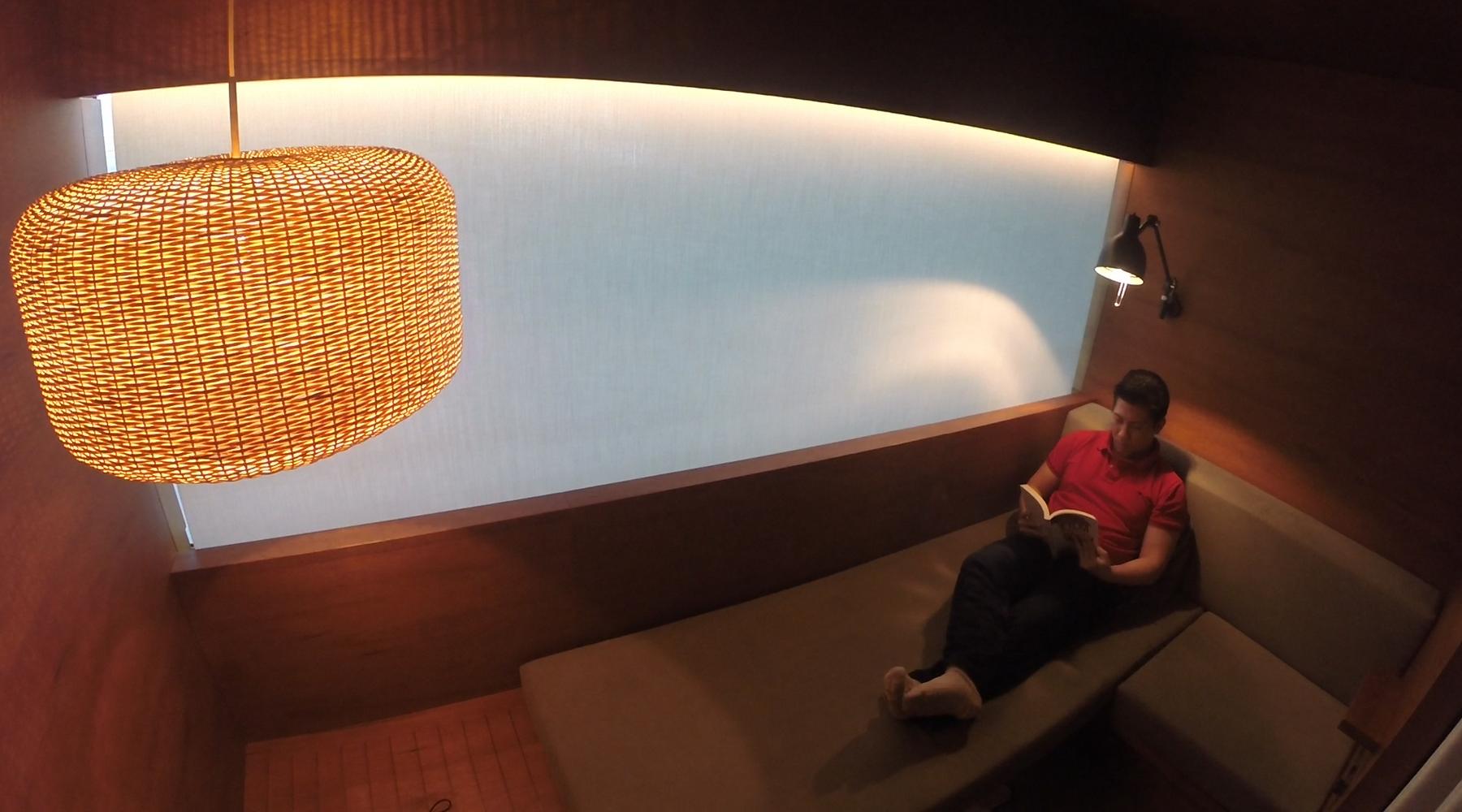 A man in a red shirt relaxes on a lounge seat, reading a book in a cozy, wood-paneled room with warm lighting.
