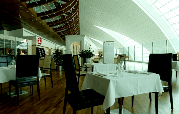 Modern airport lounge restaurant with set tables under a high, curved ceiling.
